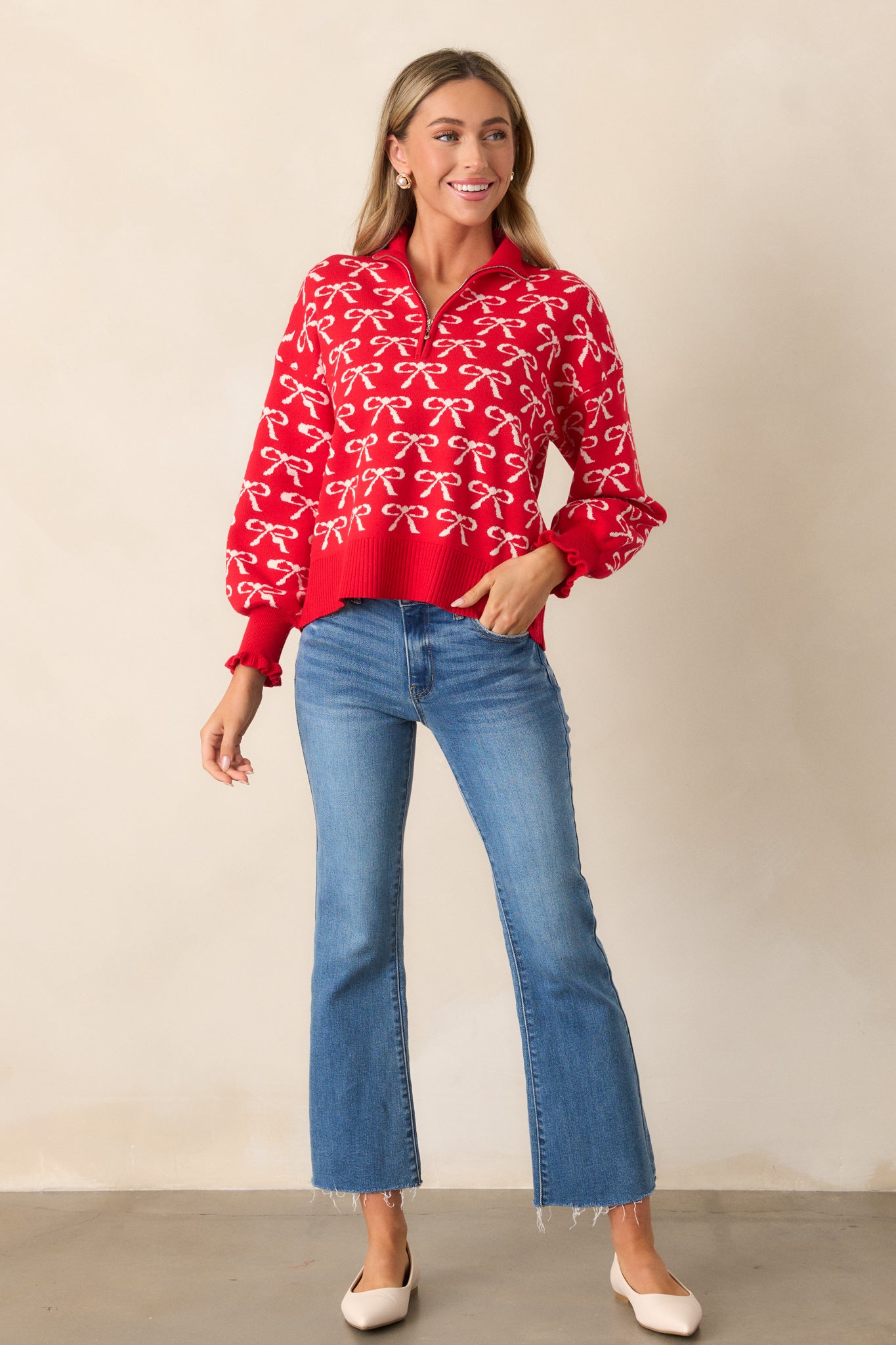 A front view of the red pullover featuring a quarter zip collared neckline, with an ivory bow print scattered throughout the fabric. The top’s ribbed hems and ruffle cuff long sleeves are visible.