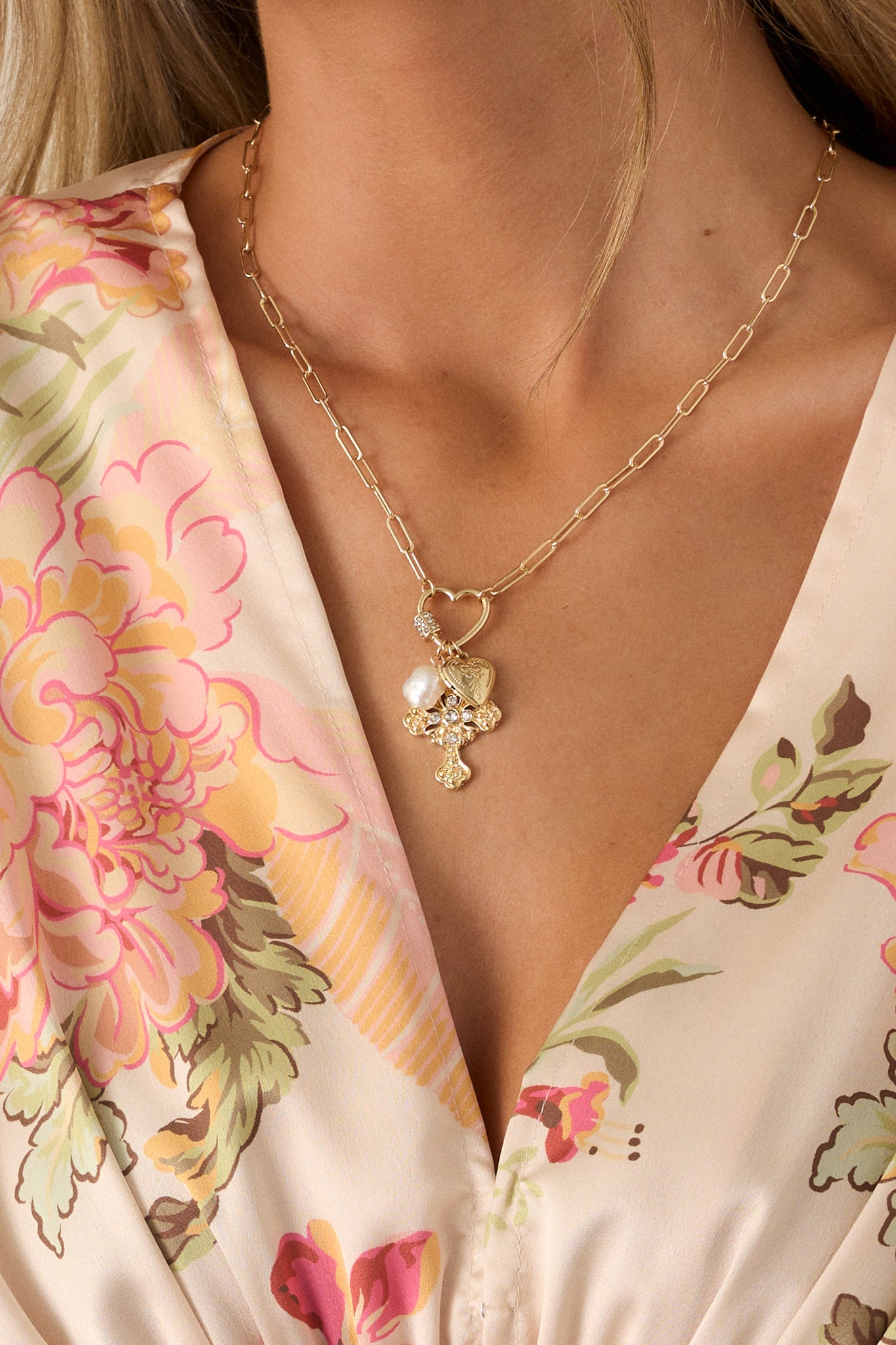 A zoomed-in view of the charms on the necklace, focusing on the pearl, heart locket, and textured cross hanging from the heart-shaped connector.