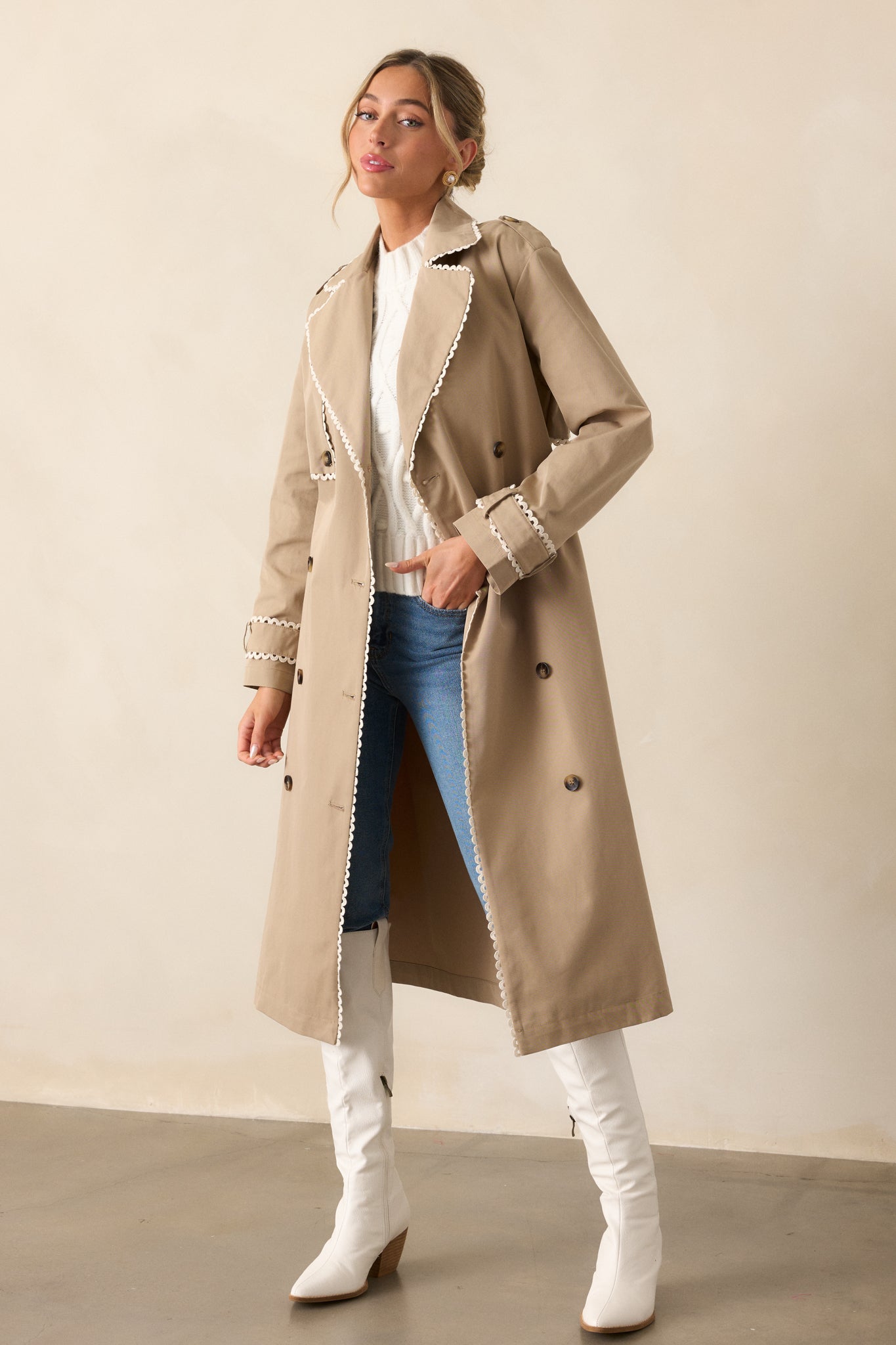  A full view of the taupe trench coat highlighting the ricrac trim detail on the sleeves .