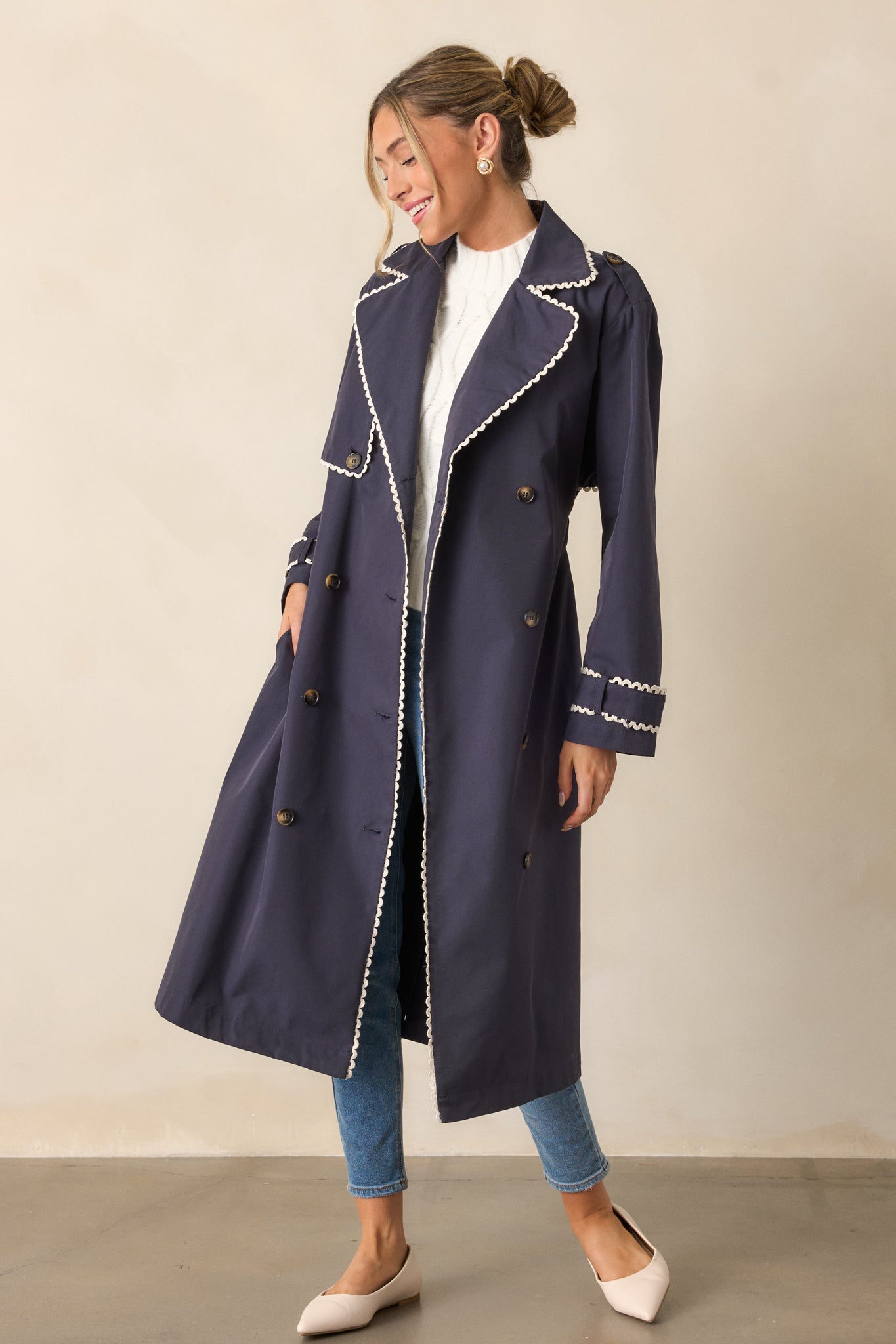 Full body view of a navy trench coat with a lapel collar, eight button detailing, and a self-tie waistband. The coat features long sleeves with button cuffs and a ricrac trim along the edges.