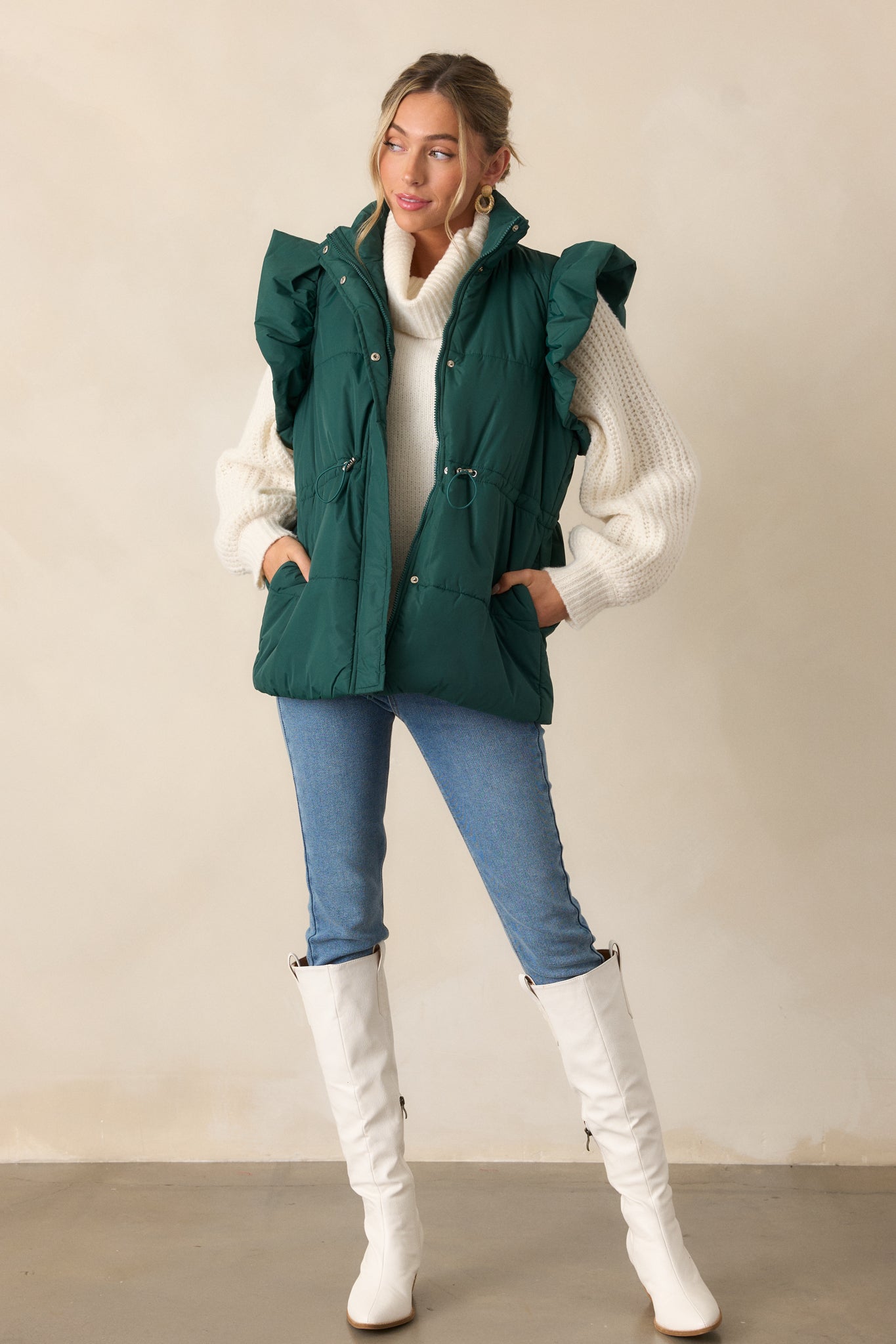 Full body view of a forest green puffer vest with a high neckline, featuring puffer line detailing, a hidden snap button and zipper front. The vest is paired with functional hip pockets and fluffy flutter sleeves.