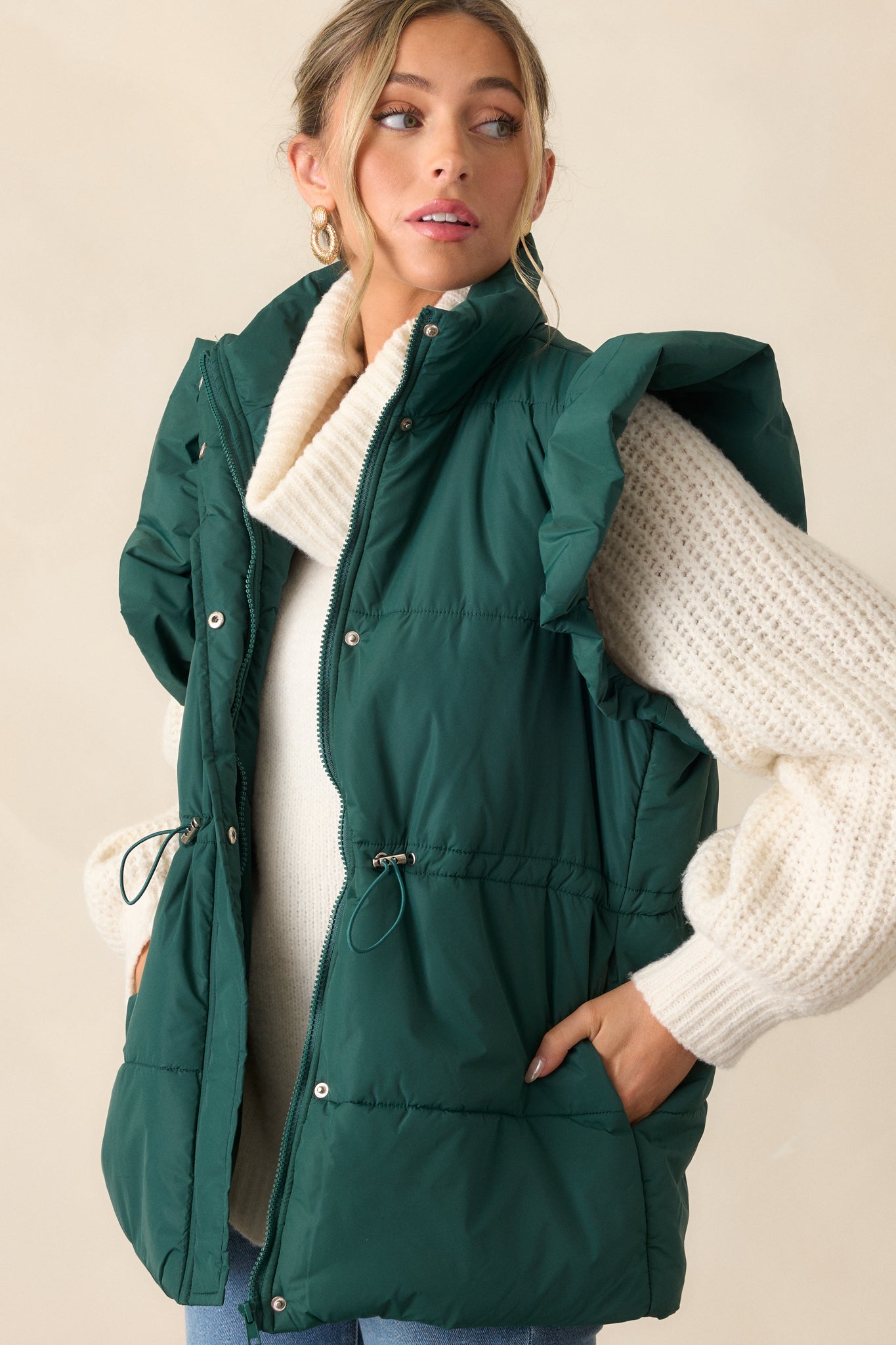 Close-up of the forest green puffer vest, focusing on the string cinch waist with silver hardware and the hidden snap button and zipper front. The texture of the puffer detailing and the smooth finish of the vest is visible.