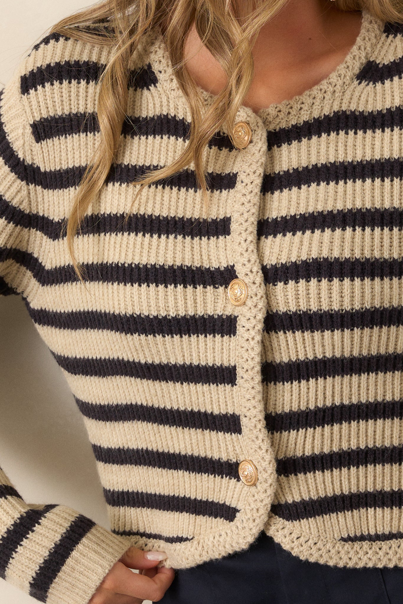 A close-up of the tan cardigan’s rounded neckline with braided knit detailing and the functional button front with gold embellished buttons. The black and tan stripe pattern is also visible, adding a stylish contrast.