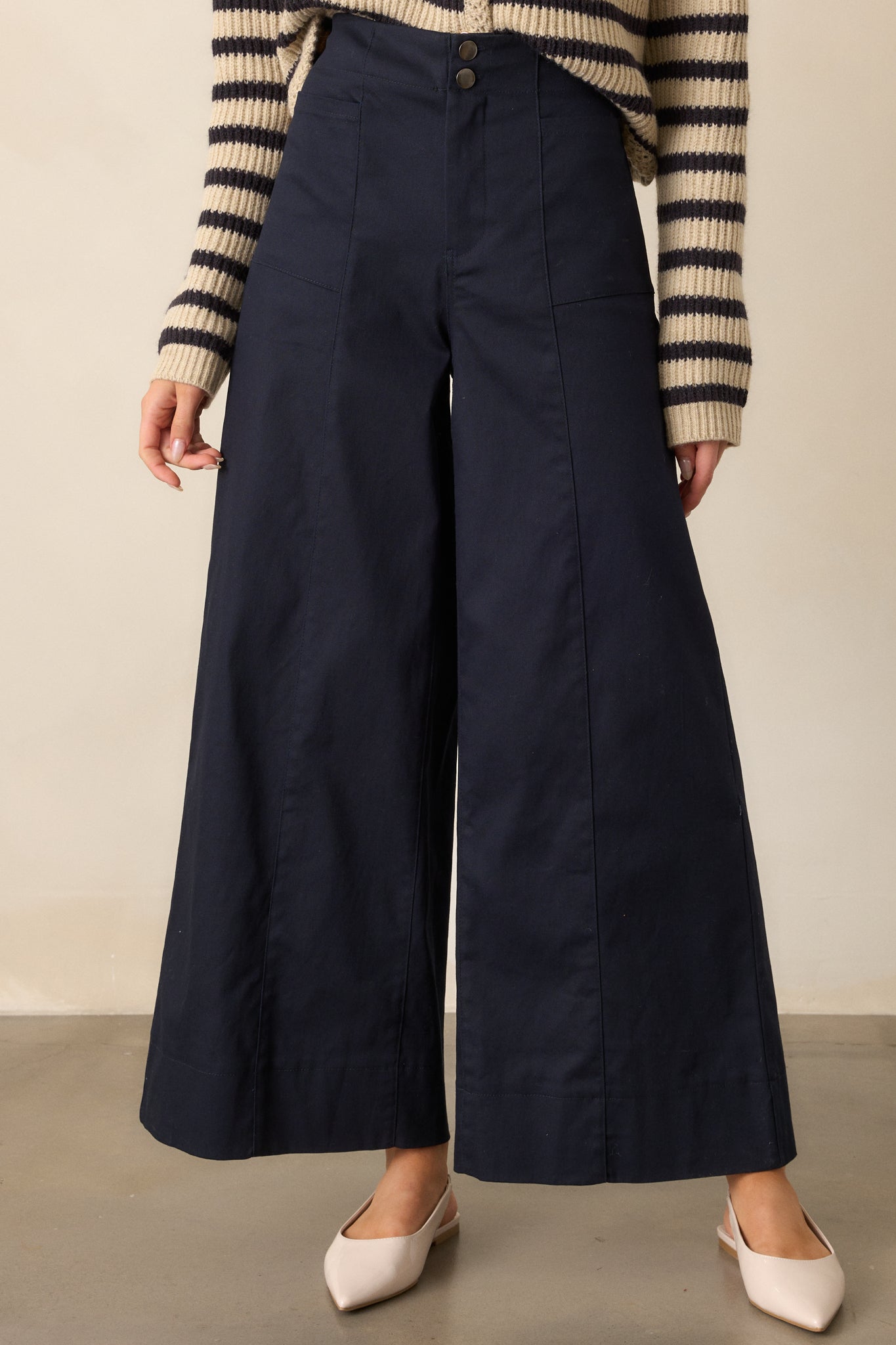 Close-up of the button and zipper closure on the high-waisted navy pants, showcasing the clean lines and structured waistband. The functional rectangular hip pockets are prominently featured, emphasizing the pants’ modern design.