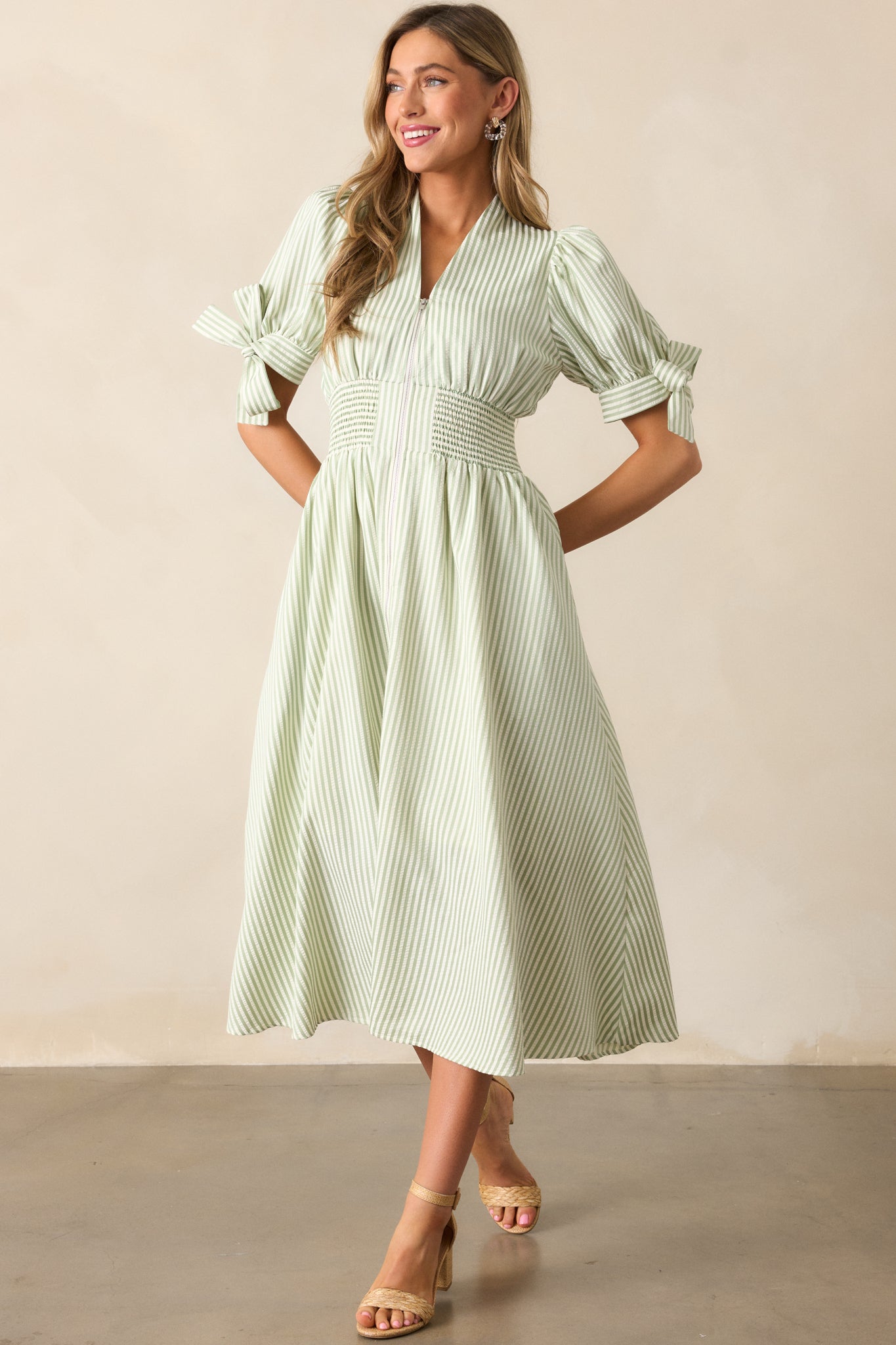 Dress in a green stripe pattern, highlighting the fitted smocked waist, front zipper, and sleeve ties, along with two functional pockets.