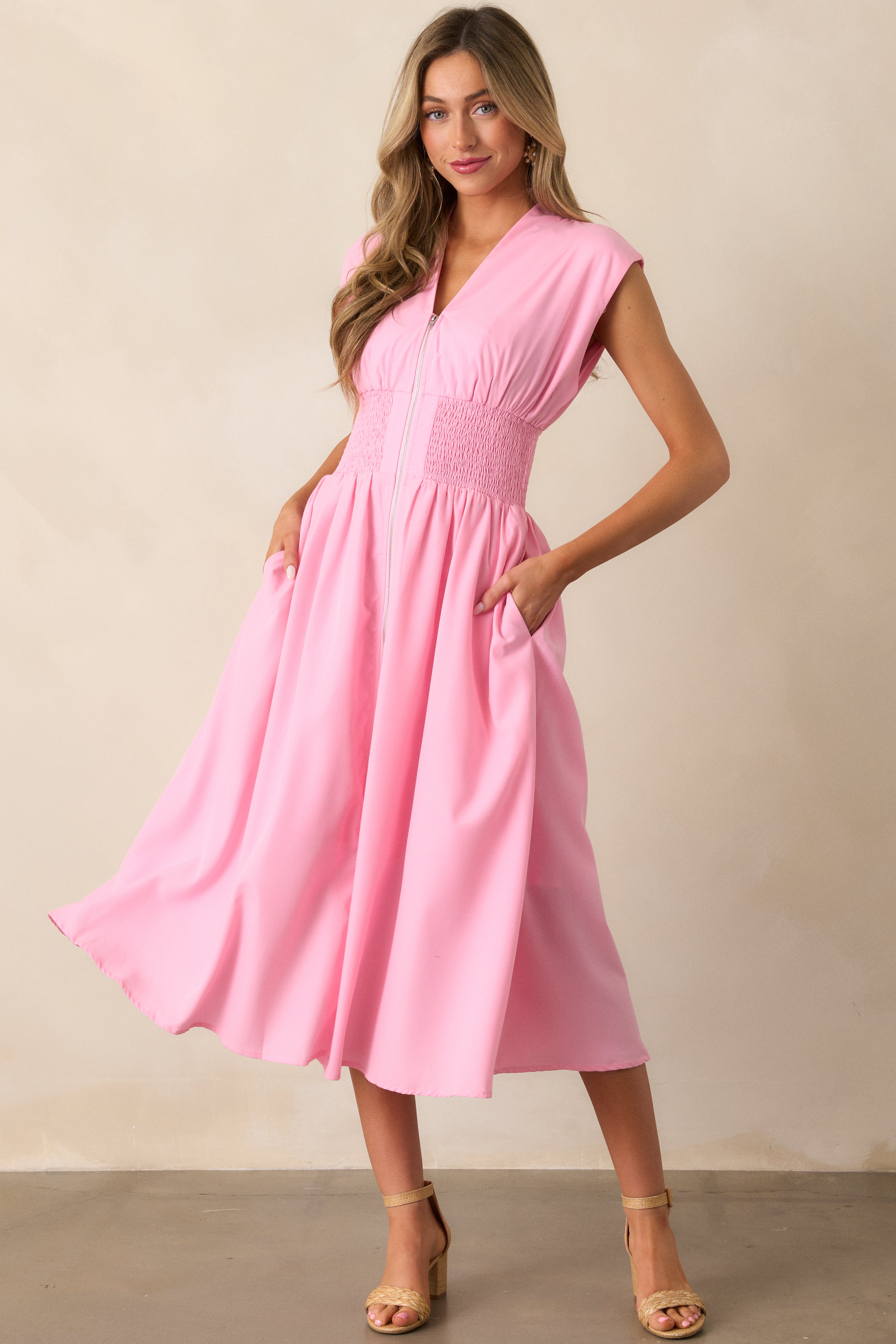 Front view of a baby pink midi dress featuring a v-neckline, padded shoulders, a full zipper front, a fully smocked waist, functional hip pockets, and a front slit.