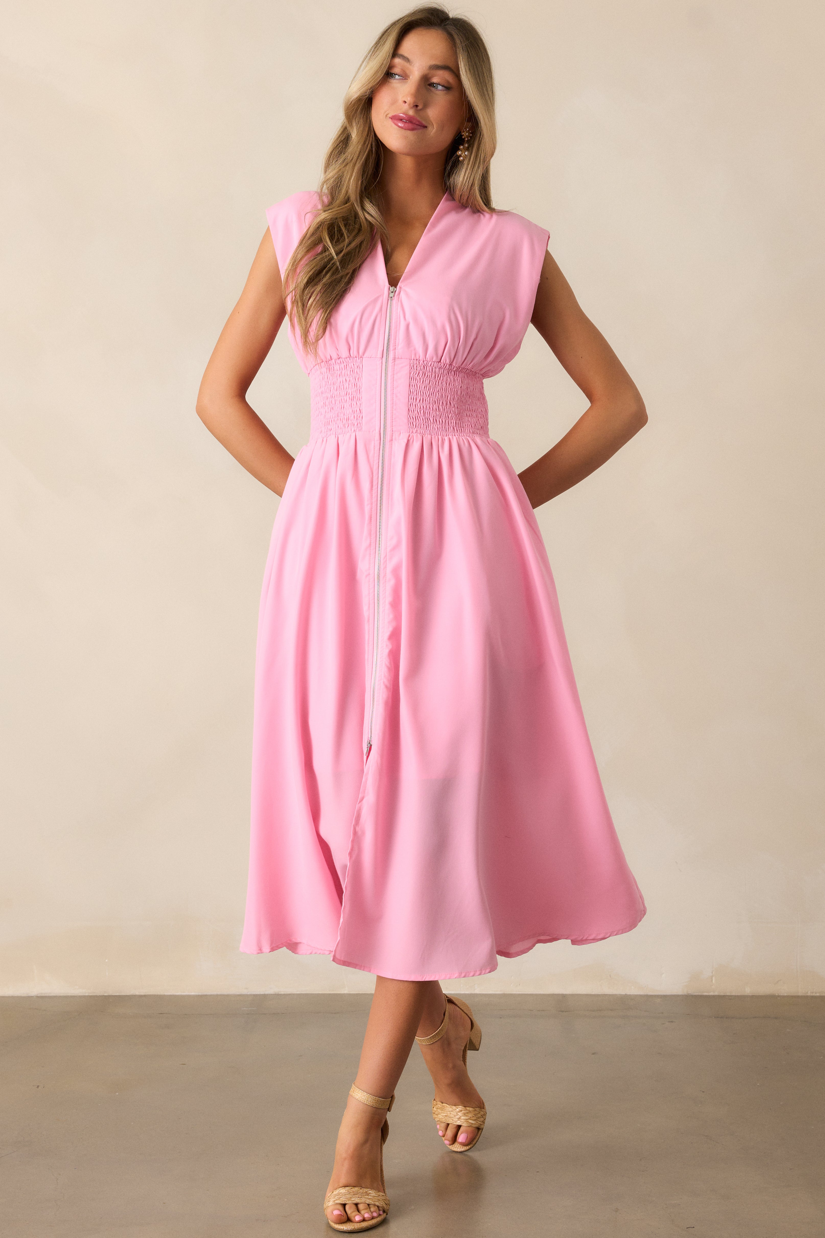 Action shot of a baby pink midi dress displaying the fit and movement, highlighting the v-neckline, padded shoulders, full zipper front, fully smocked waist, functional hip pockets, and front slit.