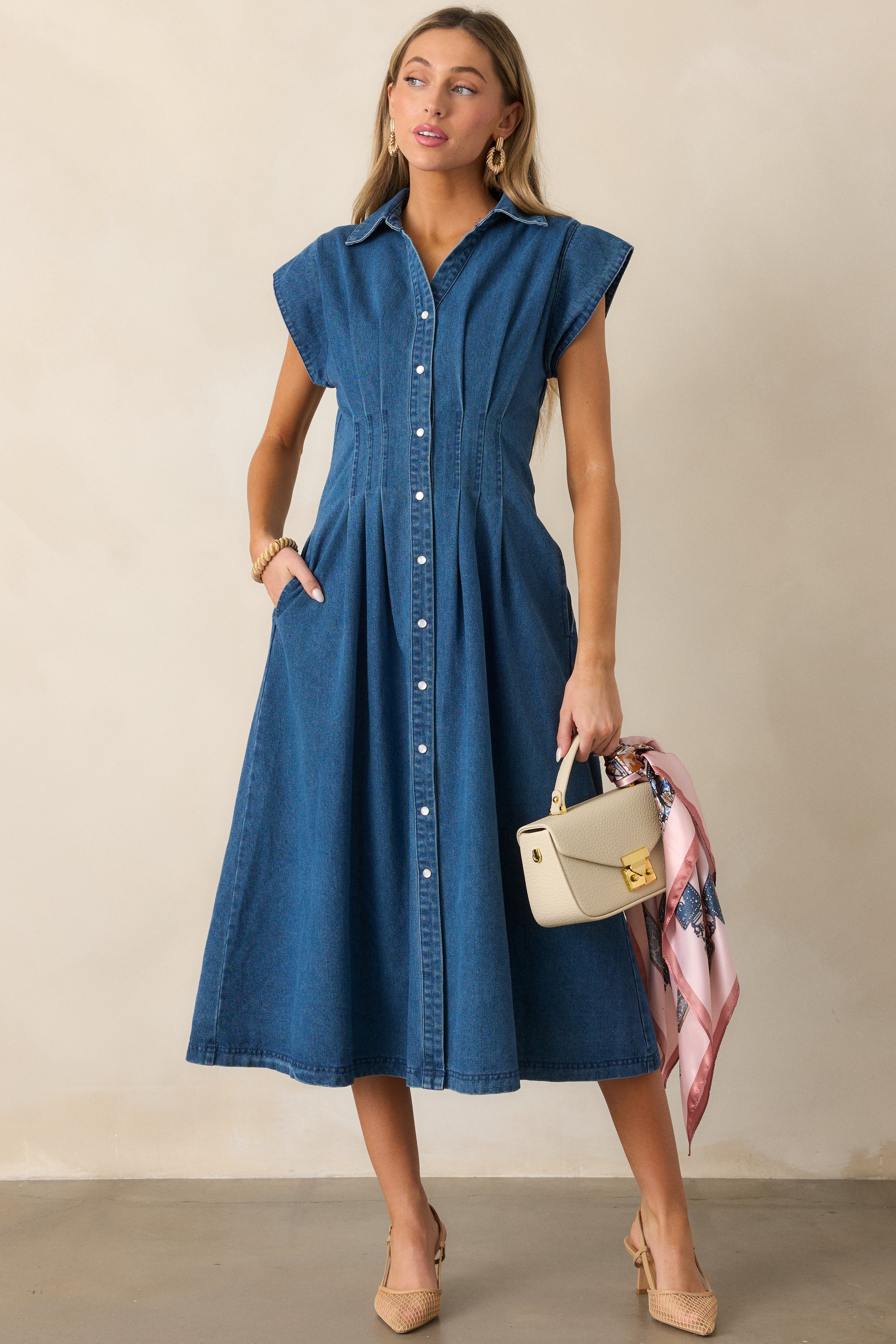 Denim dress in a dark wash featuring a button-up front with pearlized snaps, cap sleeves, and a pleated design around the waist.