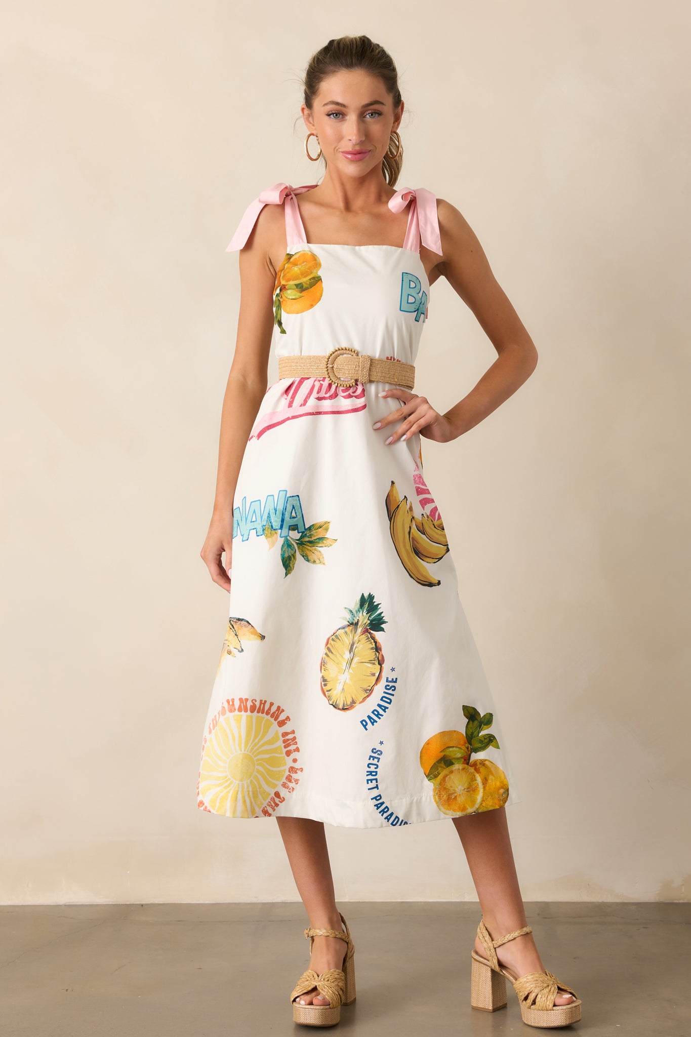 A full view of the dress showcasing its square neckline, colorful tropical fruit print, and self-tie pink ribbon straps.