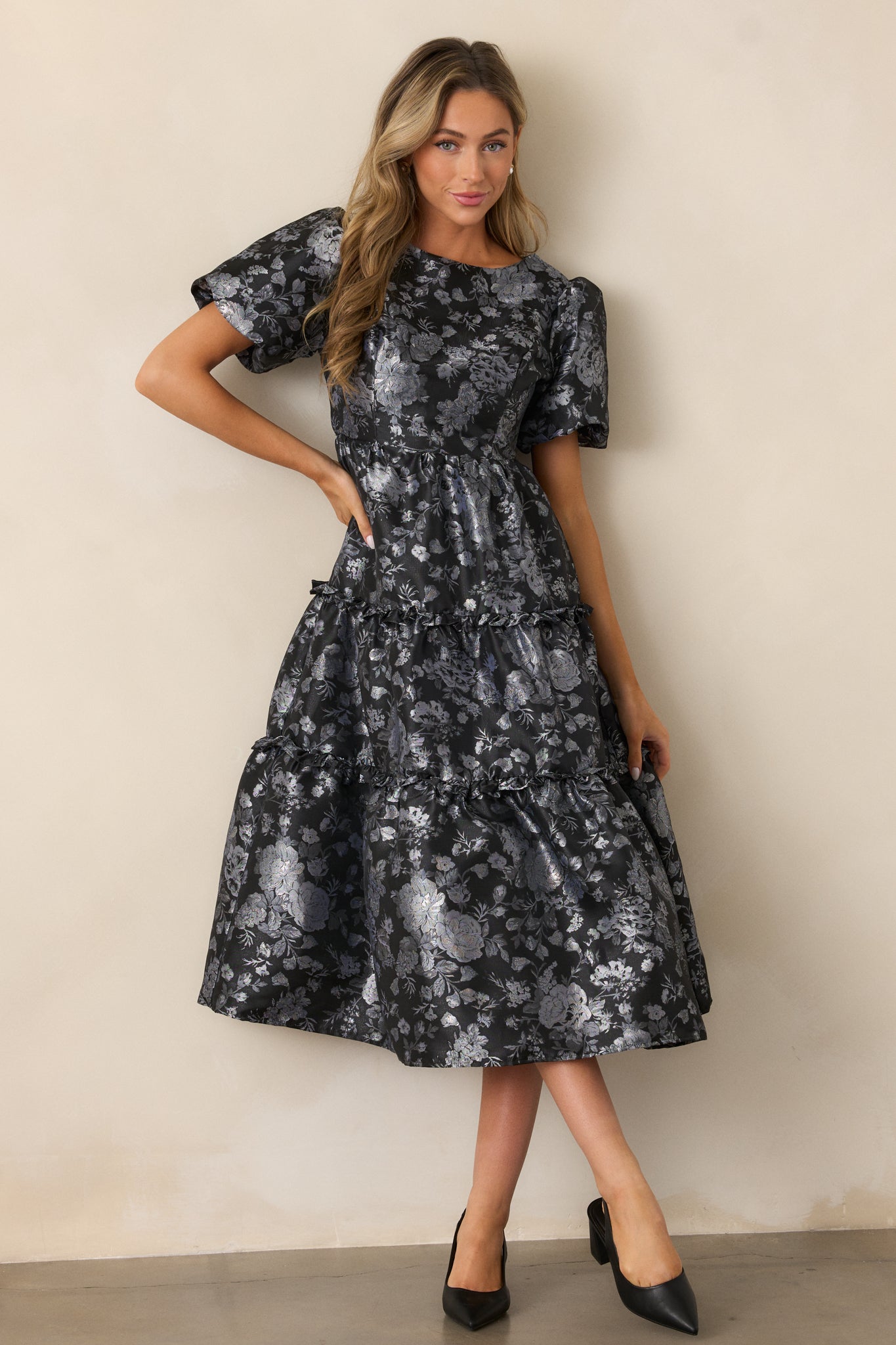 A full-length view of the black midi dress, showcasing the rounded neckline, short puff sleeves, and shiny silver floral pattern throughout.