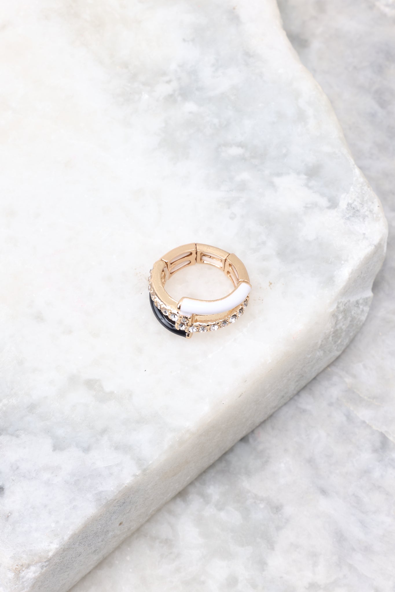 Overhead marble shot of ring that features gold hardware, black and white accents, rhinestone detailing, and elastic under the band measuring 0.75" in diameter. 