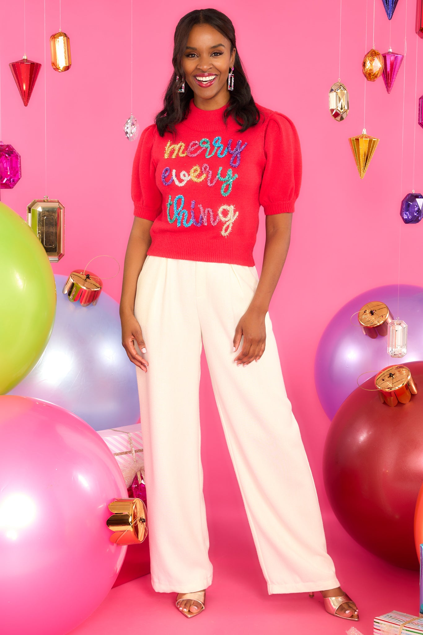 Full body view of this sweater that features a slight turtle-neck, short puff sleeves with ribbed cuffs, a ribbed hem, and multi-colored tinsel script. 