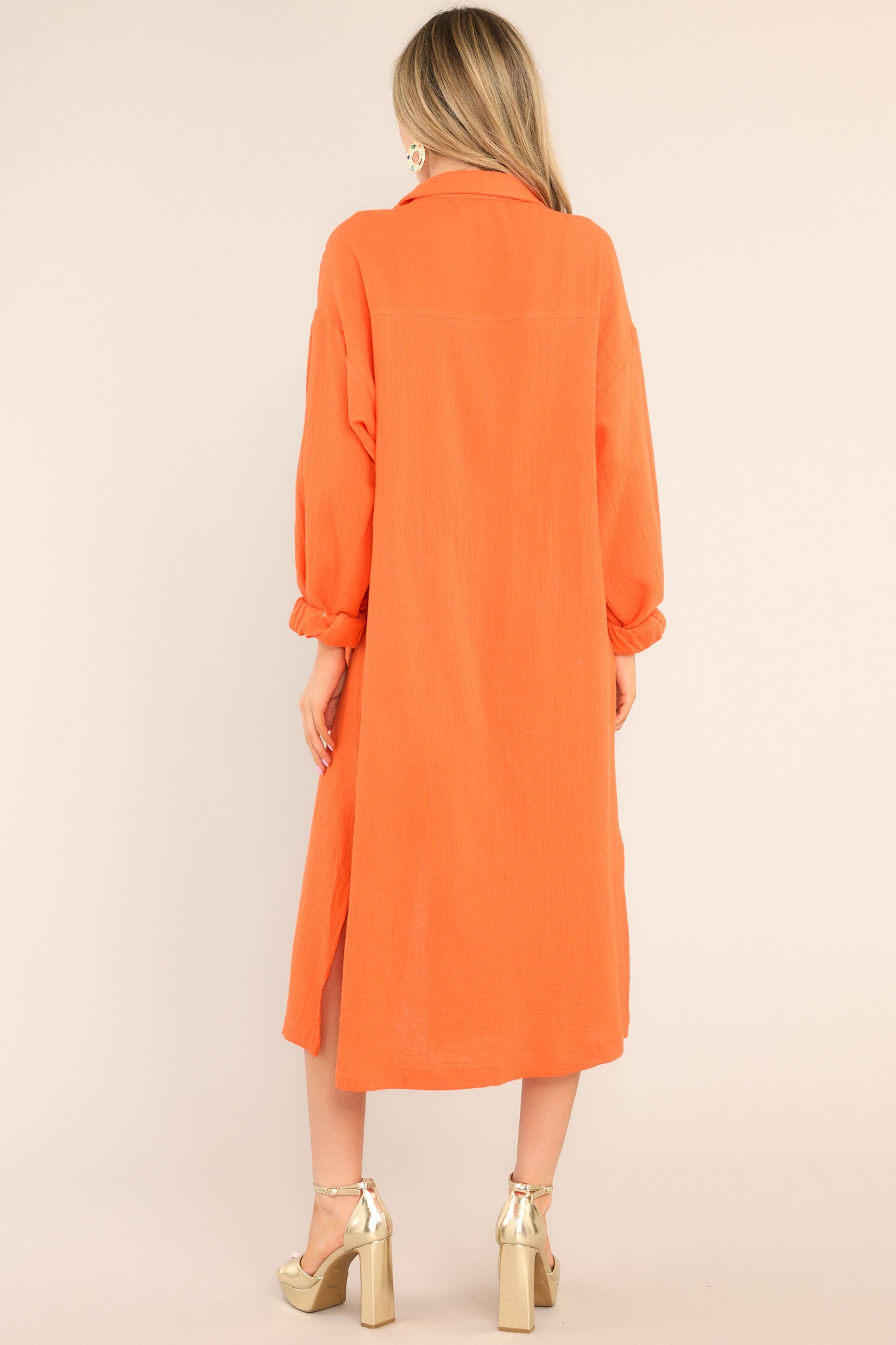 Back view of this dress that features a collared neckline, functional buttons down the front, a front pocket on the left side of the bust, long sleeves with a cuff secured by a functional button, and two slits up the bottom hemline ending just below the knee.