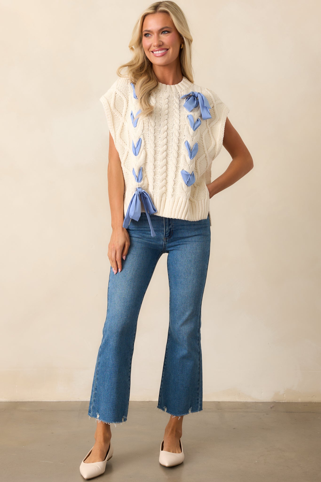 An ivory cable knit top with an oversized fit, featuring a classic crew neckline, short cap sleeves, and a delicate periwinkle ribbon woven through the front.