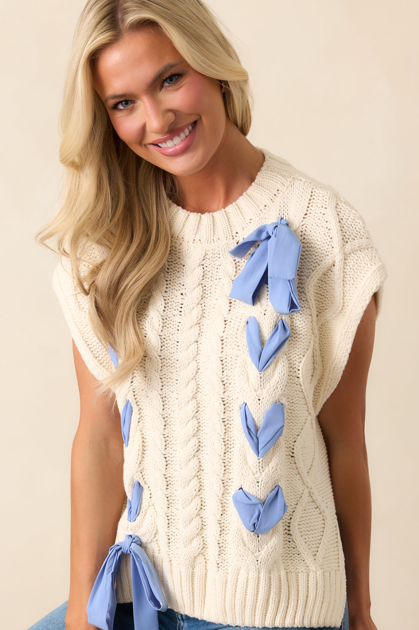 This cozy oversized ivory top showcases a textured cable knit design, ribbed hems, and a charming periwinkle ribbon detail.