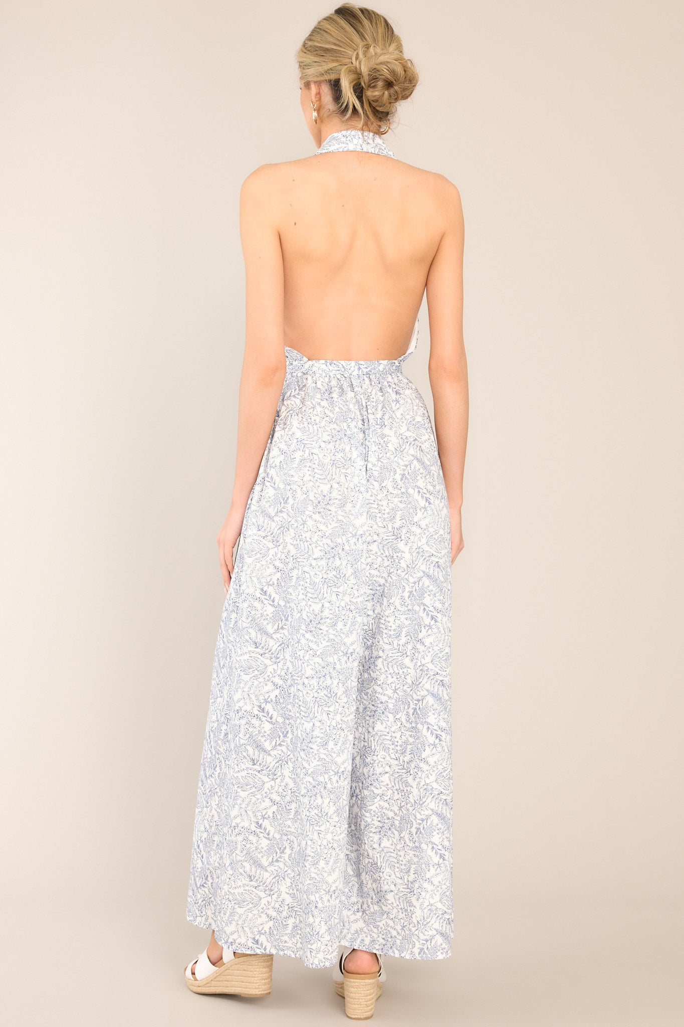 Back view of this white and navy jumpsuit that features a collared haltered v-neckline, a button front, an open back design, an elastic band at the back of the waist, belt loops, a self-tie belt, and a wide leg. It features an open back.