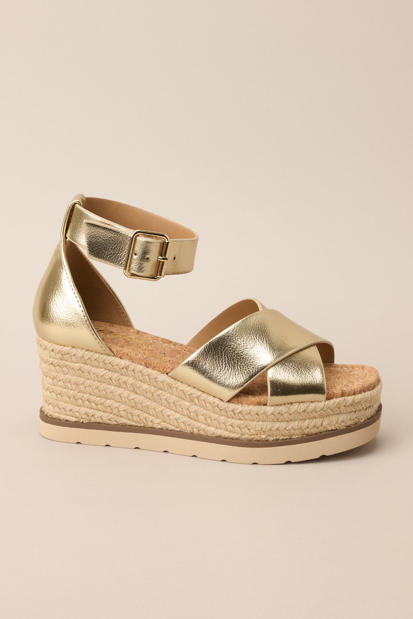 Full side view of these sandals that feature a rounded toe, gold hardware, crossed straps over the top of the foot, an ankle strap with a classic buckle closure, and an espadrille wedged platform.