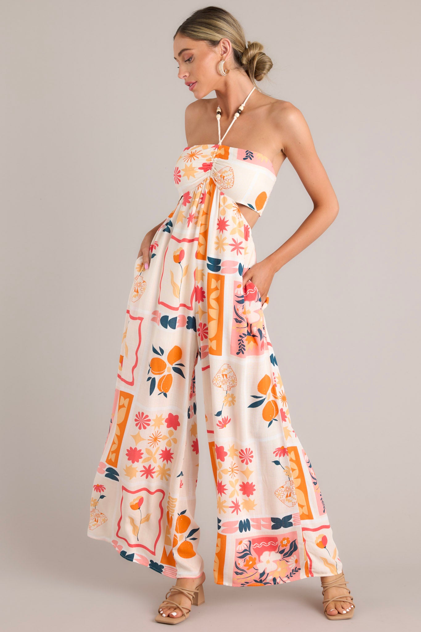 Front and side view of a patterned jumpsuit featuring a halter tassel tie neckline, an elastic waist, cutouts at the waist, functional pockets, and a smocked insert in the back of the bust.