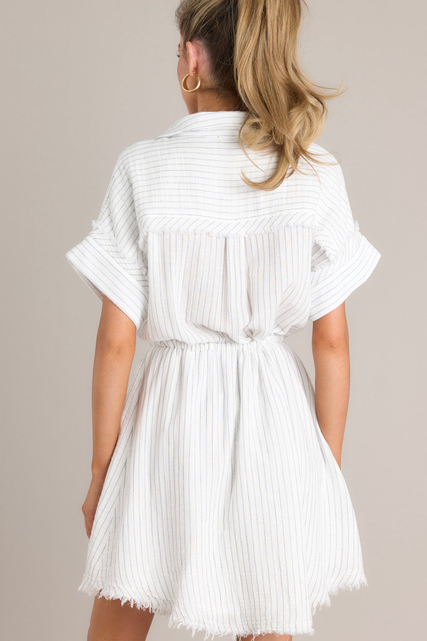 Back view of this dress that features a collared neckline, a partial button front, a self-tie drawstring waist, a raw hemline, and thick cuffed sleeves.