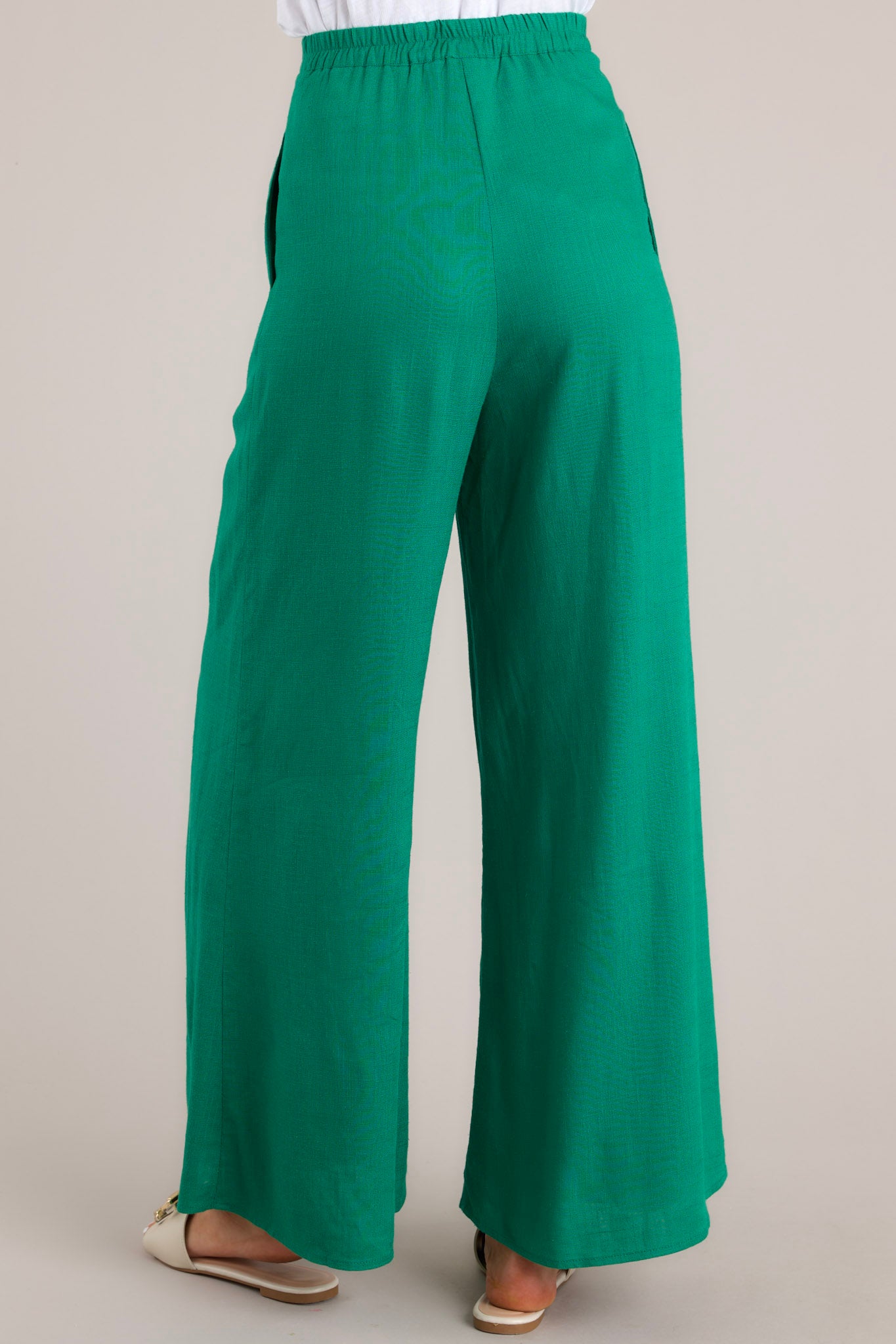 Back view of these pants that feature a high waisted design, a functional button and zipper closure, belt loops, an elastic insert at the back of the waist, hip pockets, and a wide leg.