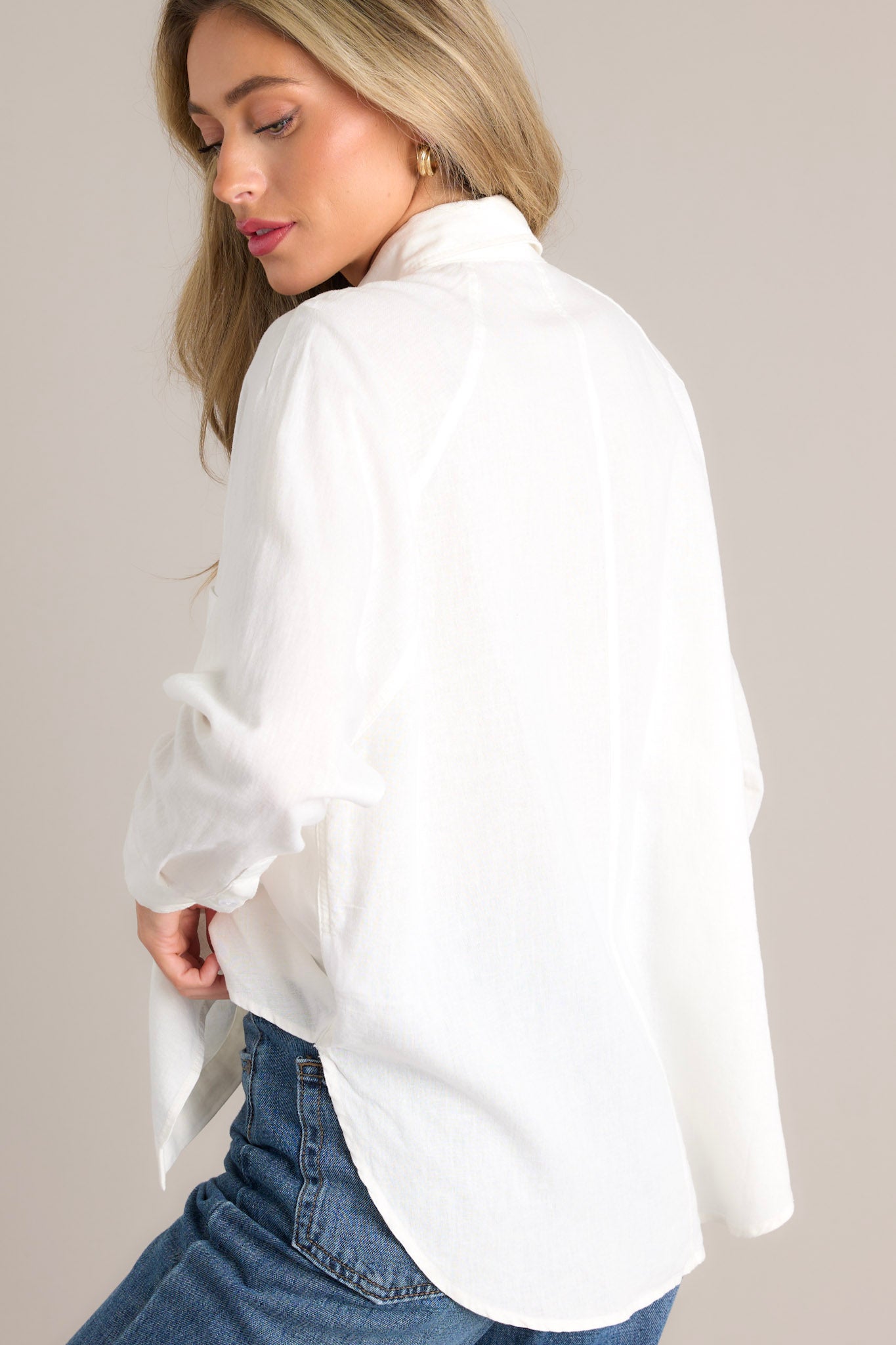 Angled back view of this white top featuring a collared neckline, a functional button front, a breast pocket, button cuffed long sleeves, and a split high low hemline.