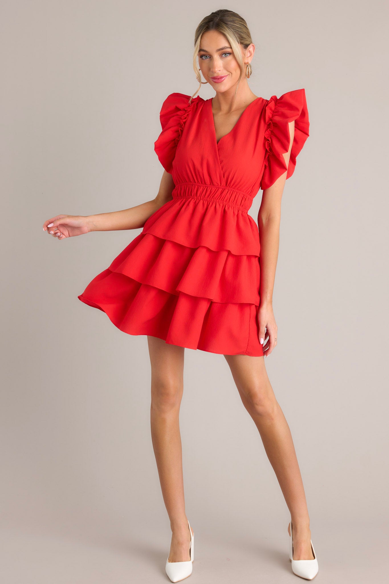 Angled full body view of this red mini dress that features a v-neckline, an elastic waist, a tiered design, and short flutter sleeves.