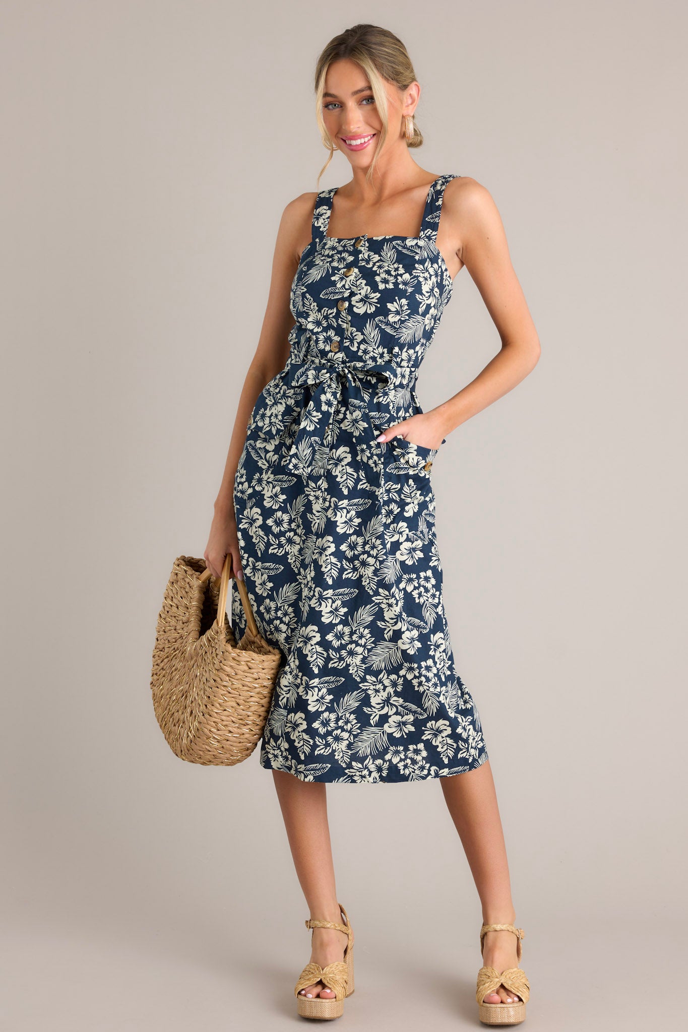 Front angled view of a navy midi dress featuring a square neckline, thick straps, functional button front bodice, self-tie waist belt with belt loops, and functional buttoned hip pockets