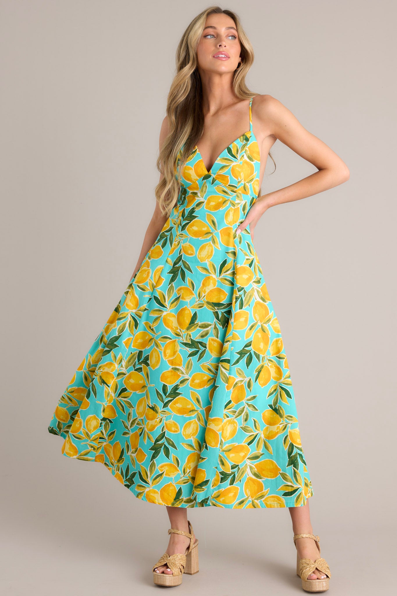 Angled front view of this aqua midi dress that features a v-neckline, thin adjustable straps with a self-tie feature, an open back with functional buttons, functional hip pockets, and, a vibrant aqua lemon print.