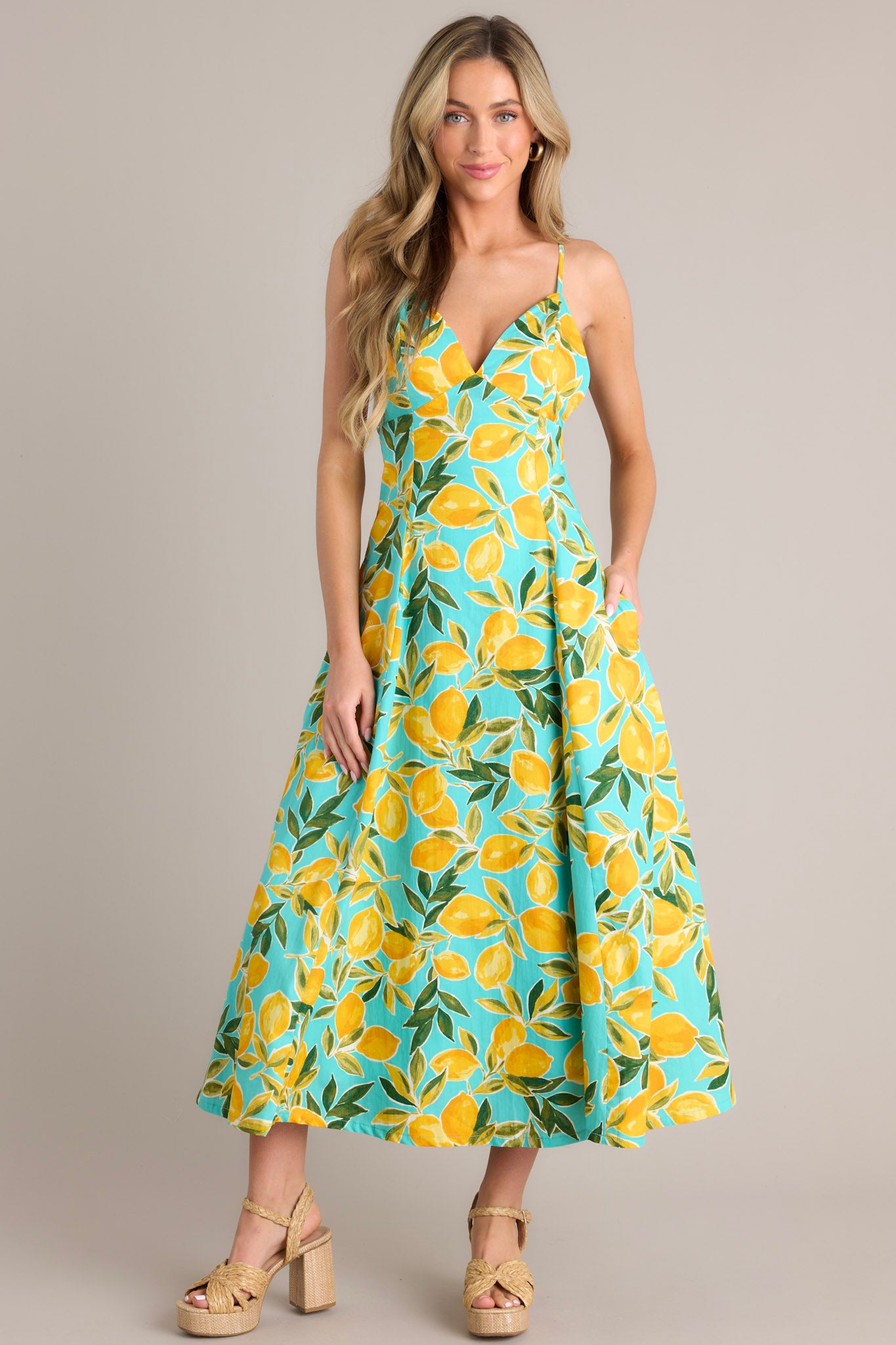 Full body view of this aqua midi dress that features a v-neckline, thin adjustable straps with a self-tie feature, an open back with functional buttons, functional hip pockets, and, a vibrant aqua lemon print.