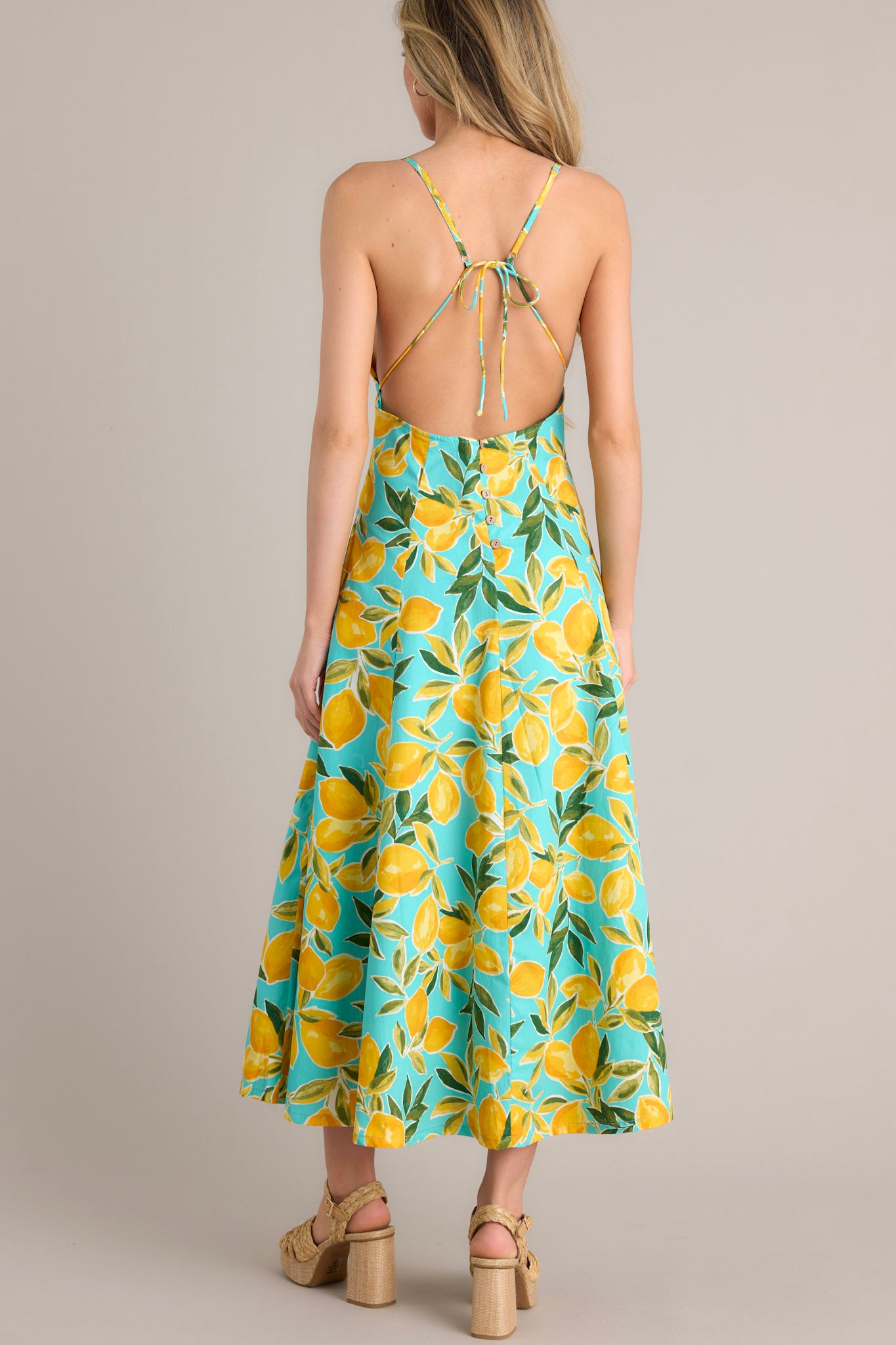 Back view of this aqua midi dress that features a v-neckline, thin adjustable straps with a self-tie feature, an open back with functional buttons, functional hip pockets, and, a vibrant aqua lemon print.