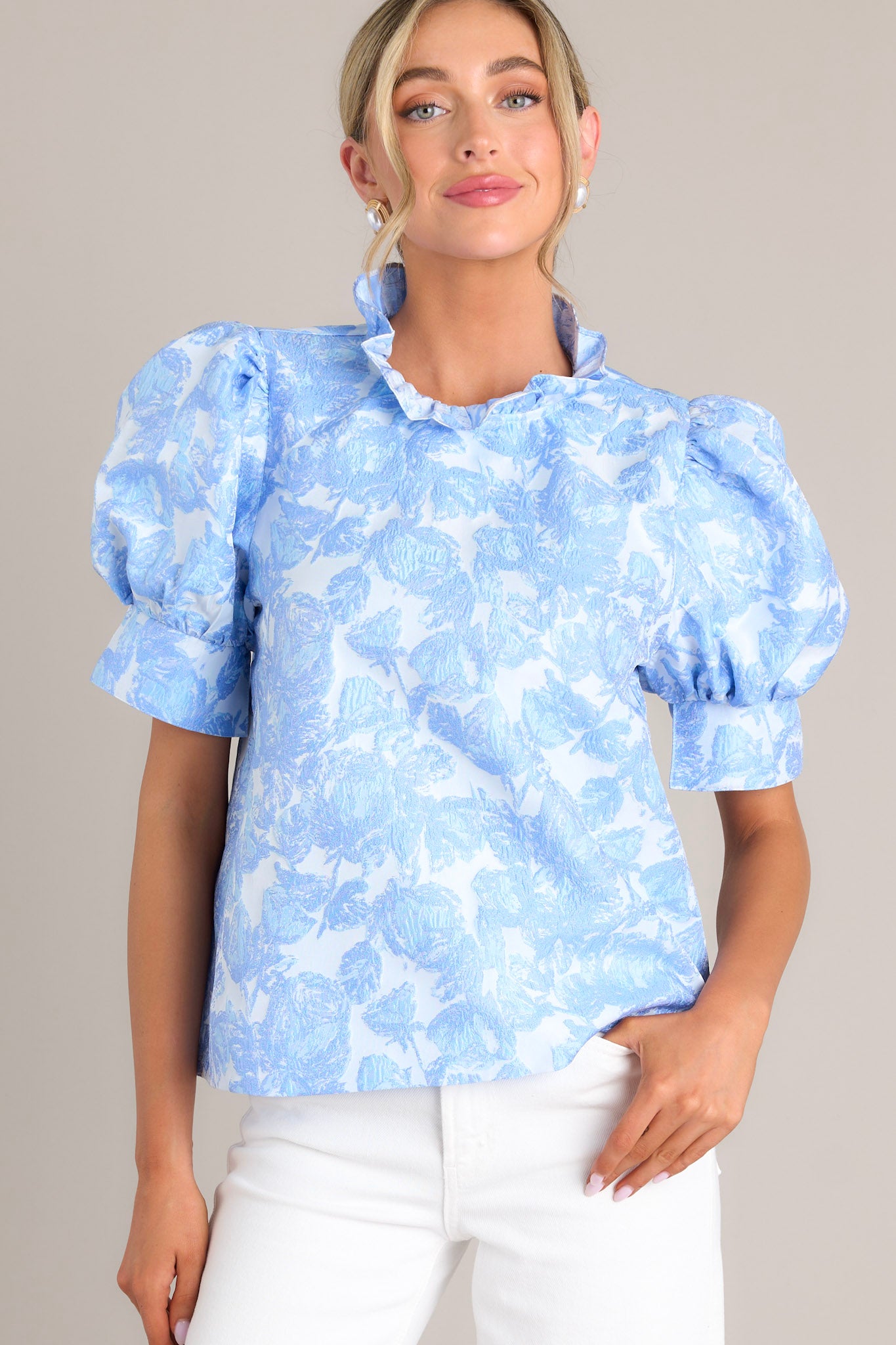 Full front view of this top that features a ruffled high neckline, functional buttons down the back, a textured material, and thick cuffed puff sleeves.