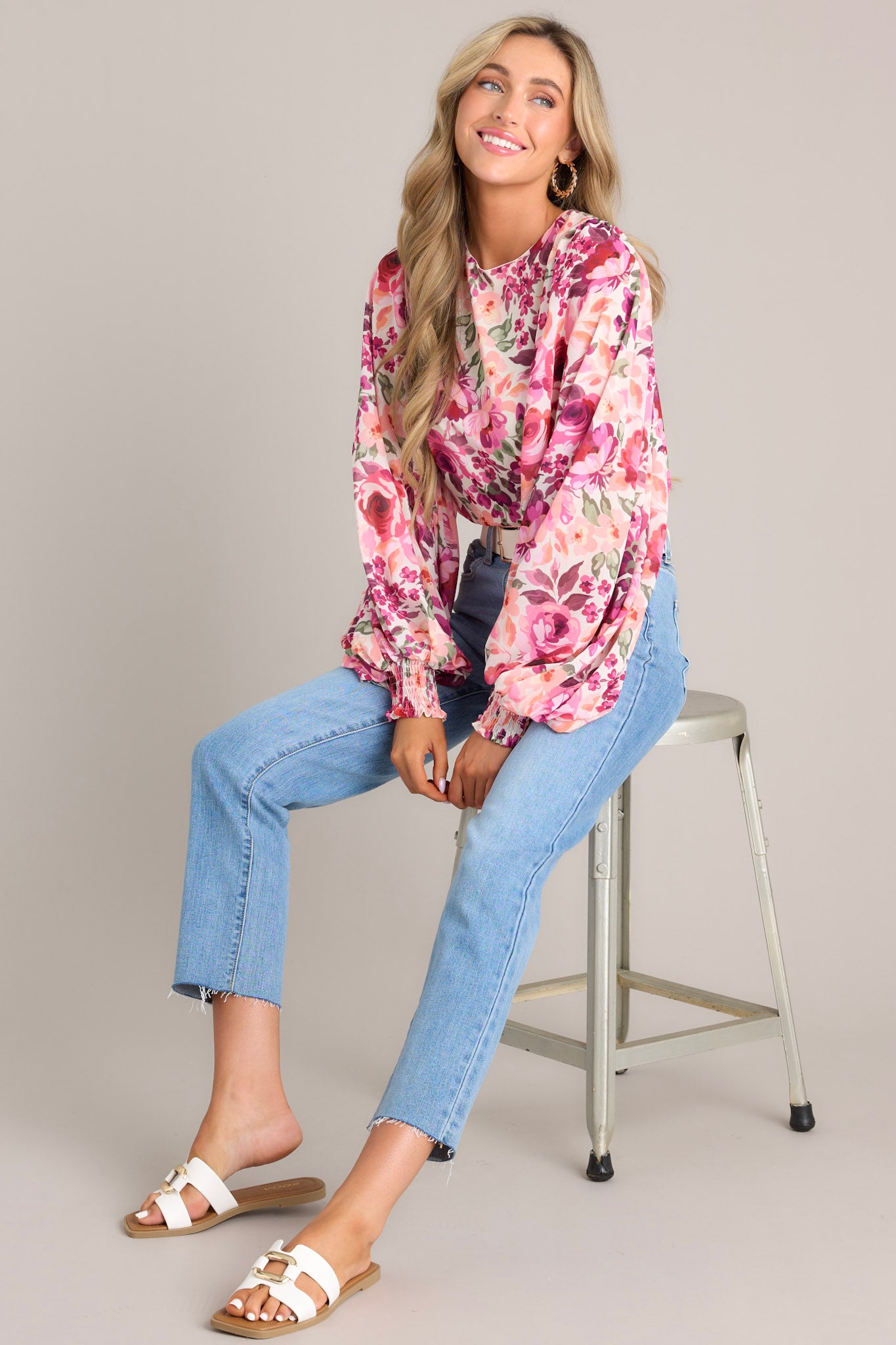 Seated Full length view of a top with a round neckline, a keyhole cutout at the back of the neck with a button closure, long sheer sleeves with smocked cuffs, and a vibrant floral pattern throughout