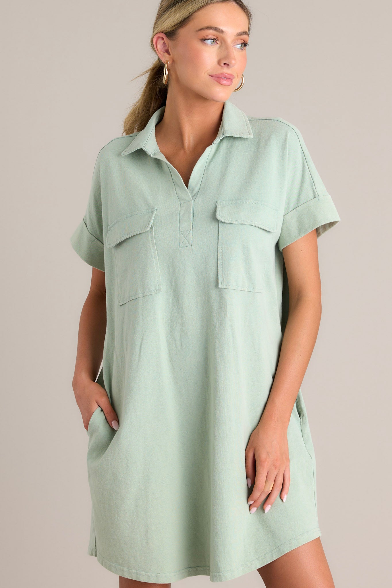 Angled view of a sage green mini dress featuring a collared neckline, functional breast and hip pockets, relaxed fit, and cuffed short sleeves.