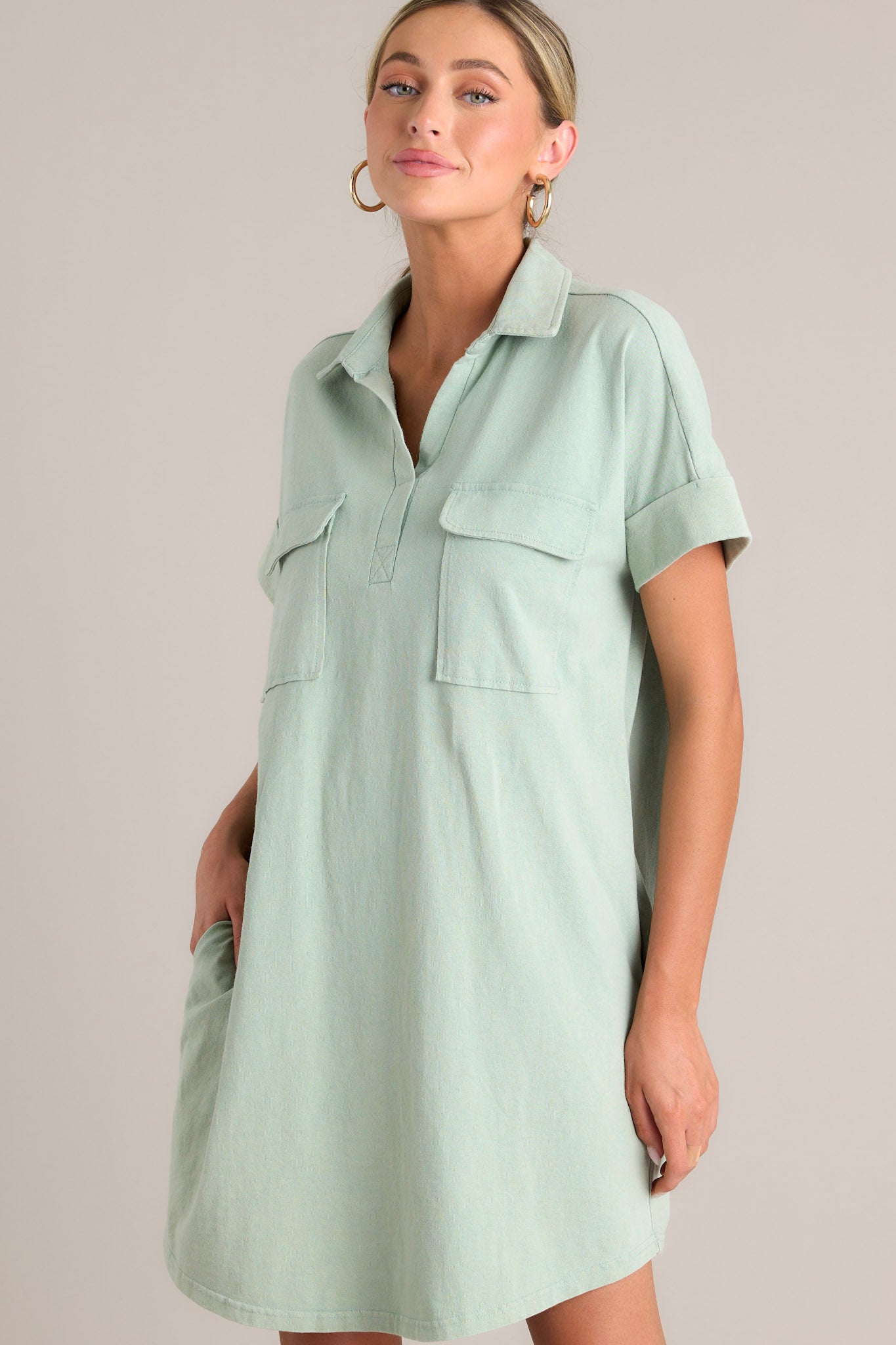 Action shot of a sage green mini dress with a collared neckline, functional breast and hip pockets, relaxed fit, and cuffed short sleeves, highlighting the fit and design.