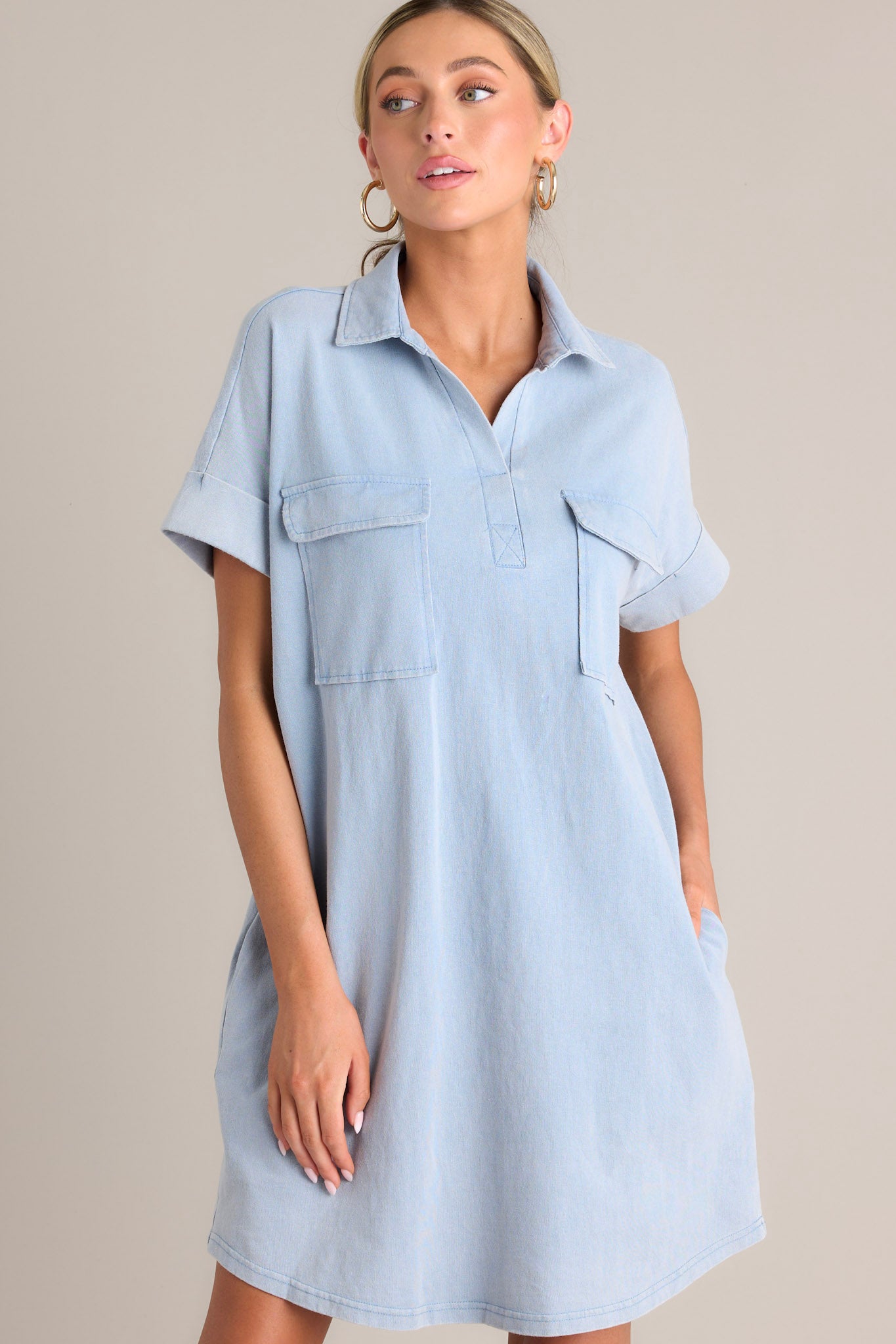 Angled front view of this light chambray mini dress that features a collared neckline, functional breast & hip pockets, a relaxed fit, and cuffed short sleeves.