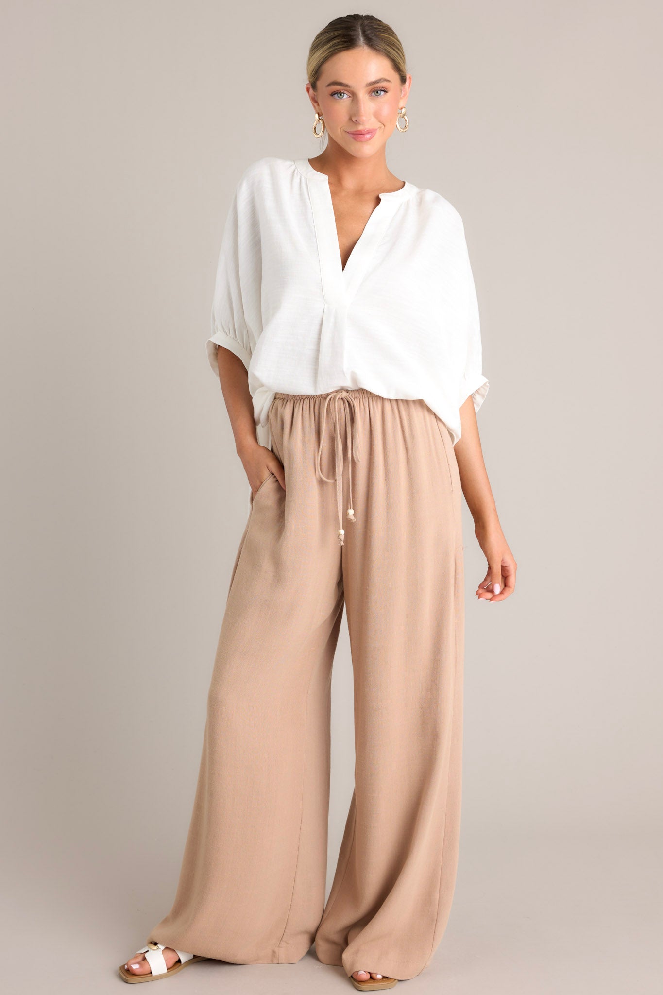 Full body view of these tan pants that feature a high waisted design, a thin elastic waistband, a self-tie drawstring, functional hip pockets, and a wide leg.