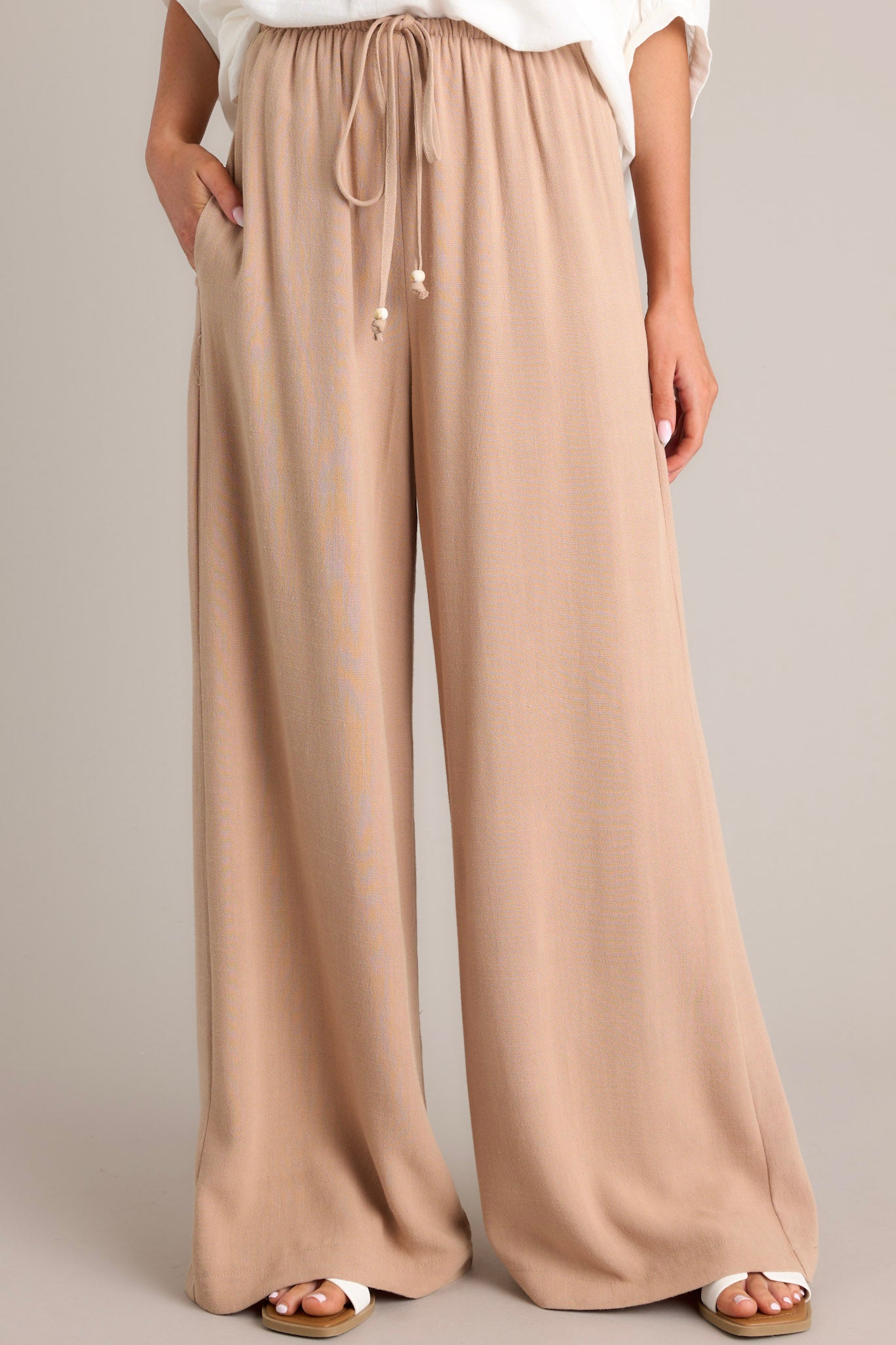 Front view of these tan pants that feature a high waisted design, a thin elastic waistband, a self-tie drawstring, functional hip pockets, and a wide leg.