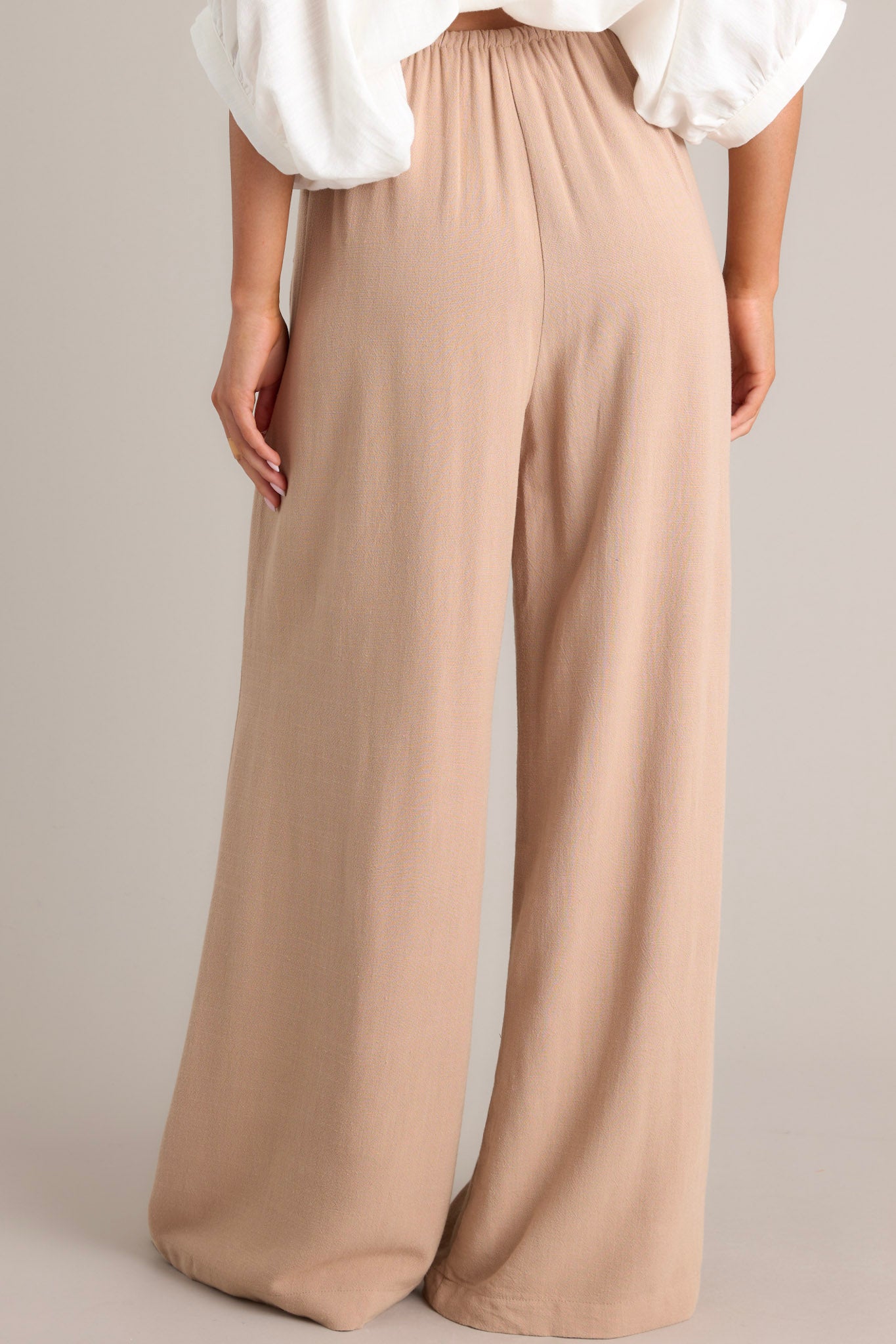 Back view of these tan high-waisted pants with thin elastic waistband, self-tie drawstring, functional hip pockets, and wide leg.