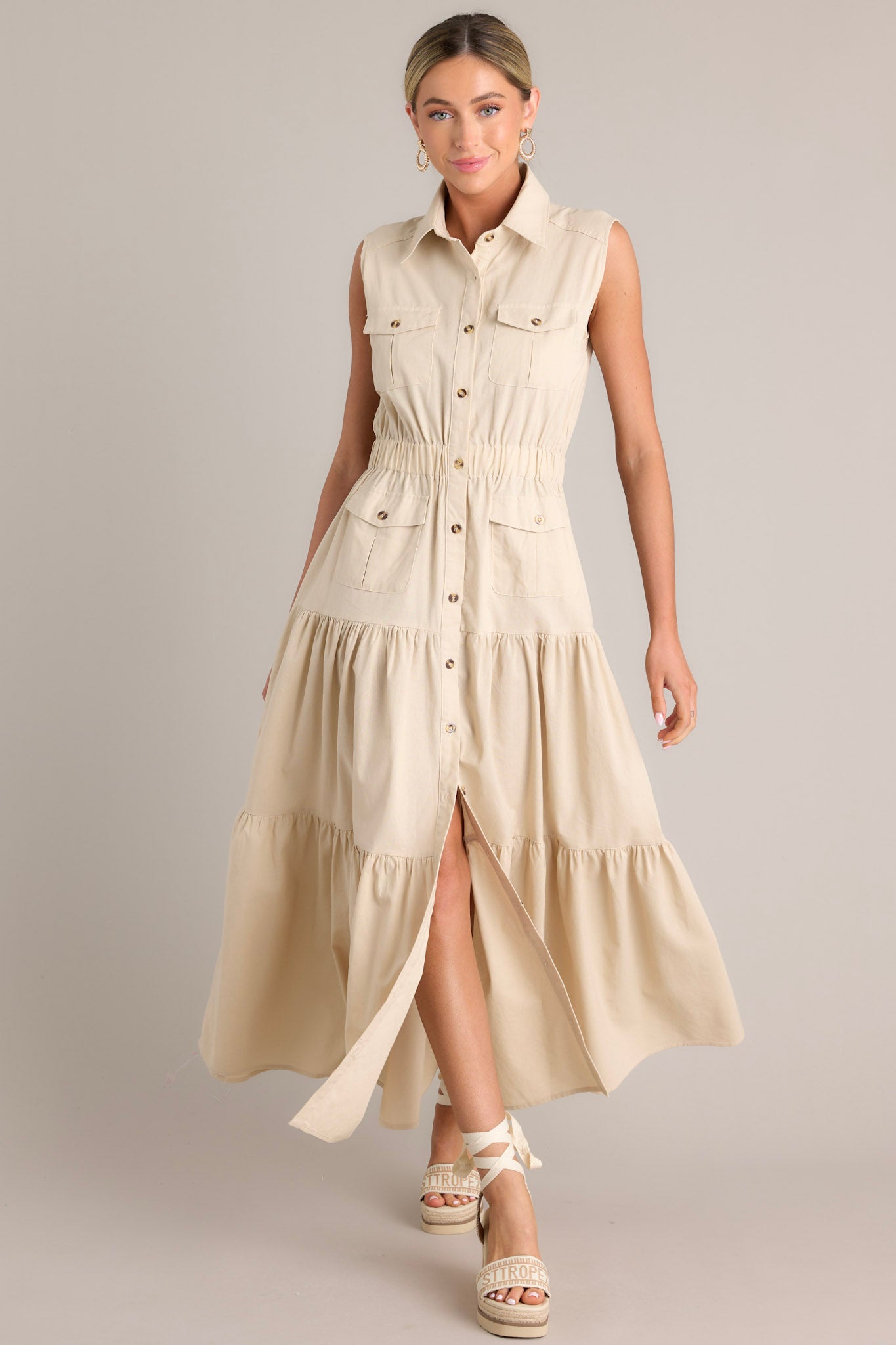 Full body view of this beige maxi dress that features a collared neckline, a full button front, buttoned breast pockets, a thick elastic waistband, buttoned hip pockets, and a tiered design.