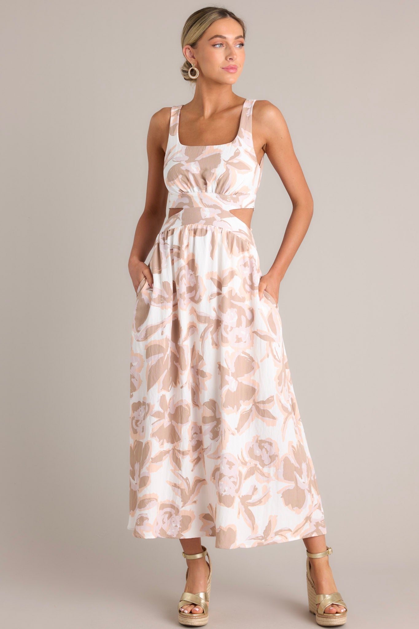 Front angled view of a white maxi dress featuring a square neckline, thick straps, elastic back insert, self-tie back feature, waist cutouts with a smocked back insert, functional hip pockets, and an all-over floral pattern
