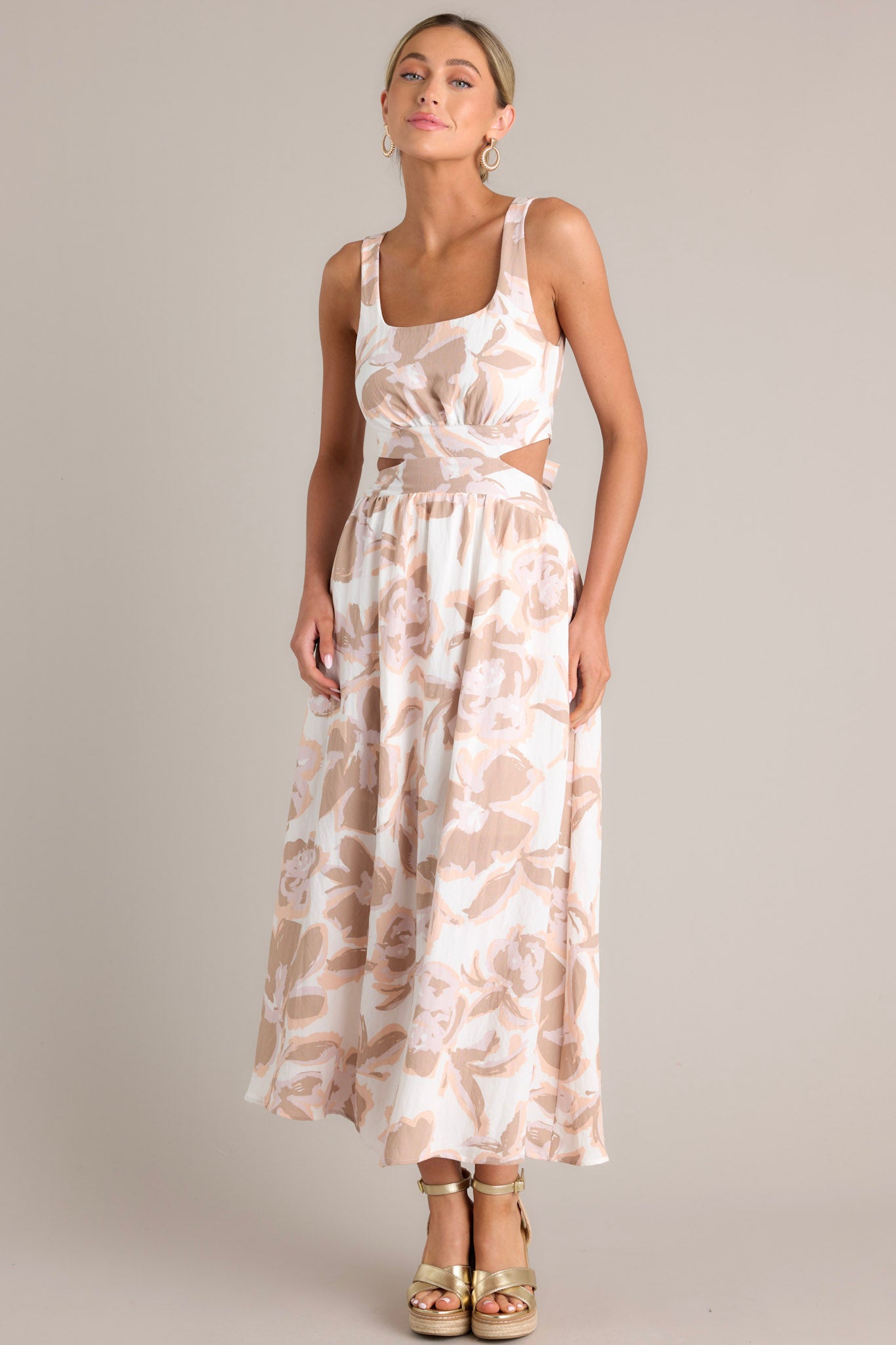 Full length view of a white maxi dress featuring a square neckline, thick straps, elastic back insert, self-tie back feature, waist cutouts with a smocked back insert, functional hip pockets, and an all-over floral pattern.