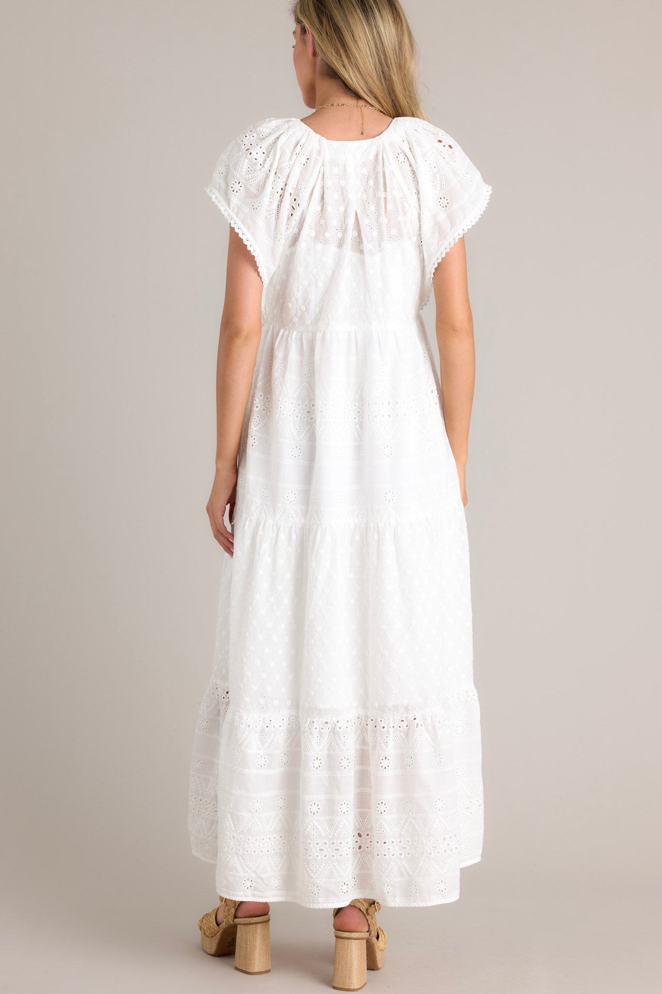 Back view of this white maxi dress with v-neckline, removable slip, thin elastic waistband, hip pockets, eyelet detailing, and faux pearl trimmed sleeves.