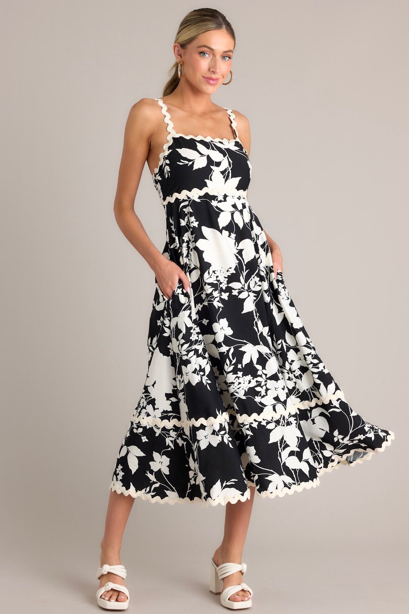 Angled full body view of  this black floral midi dress that features a square neckline, thin ricrac straps, a fully smocked back, ricrac detailing, functional hip pockets, and a ricrac hemline.