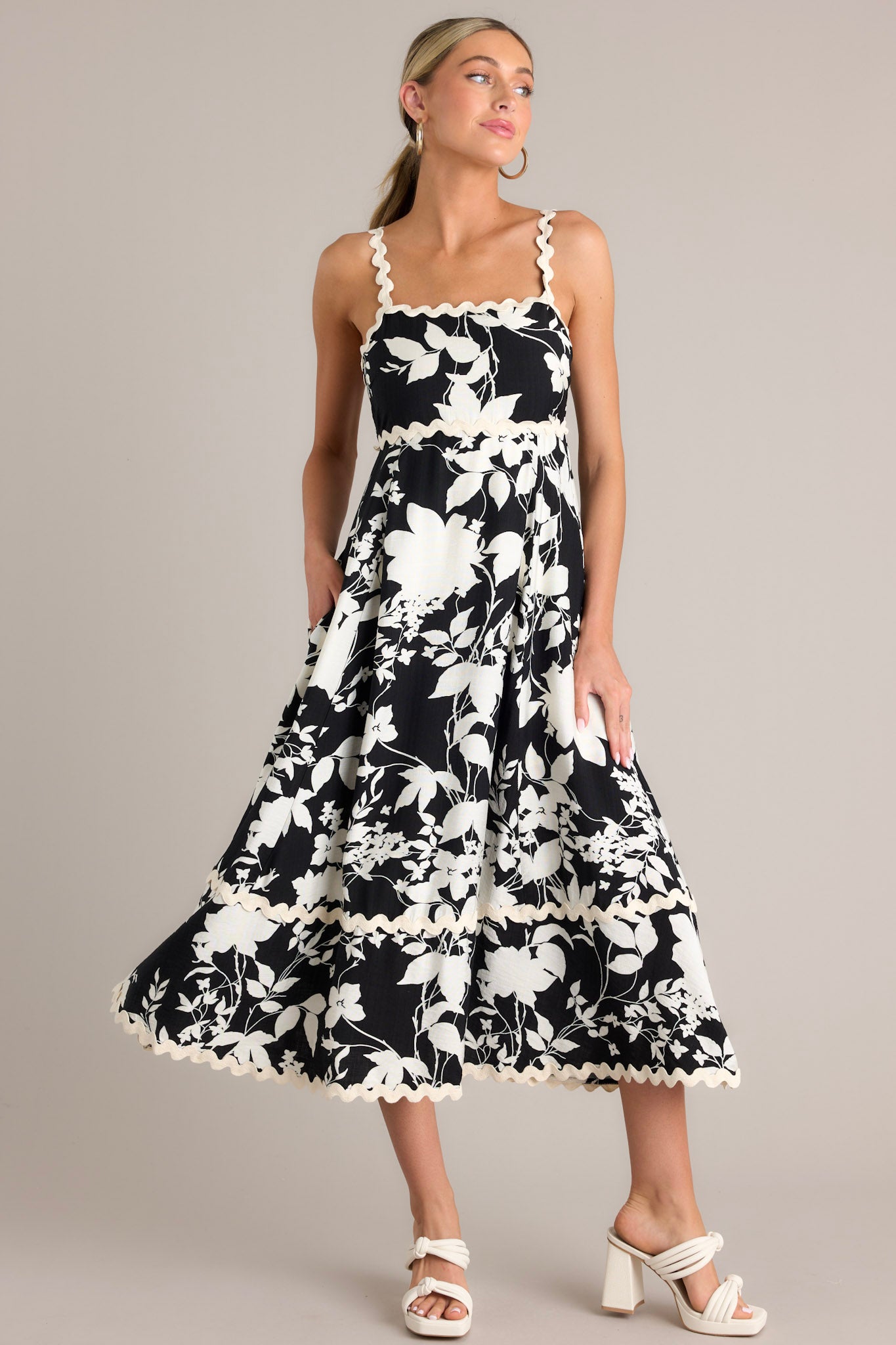 This black floral midi dress features a square neckline, thin ricrac straps, a fully smocked back, ricrac detailing, functional hip pockets, and a ricrac hemline.