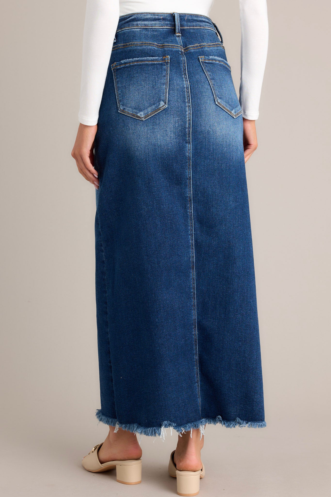 Back view of this dark wash denim midi skirt features a high waisted design, belt loops, a zipper and button closure, functional pockets, a 15" slit up the center, and raw hem detailing.