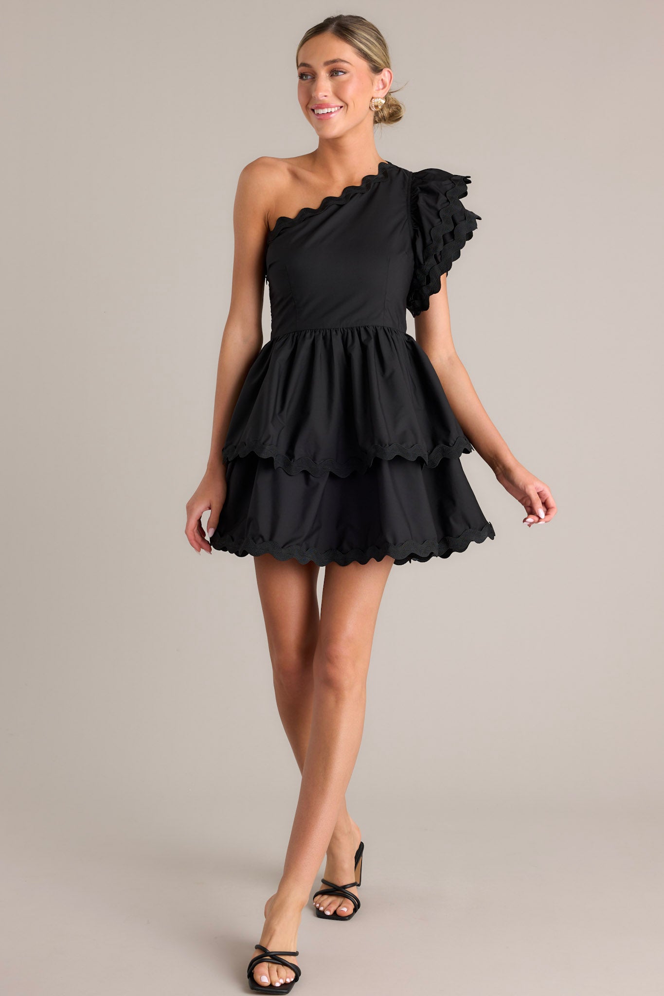 Full body view of this black romper that features rick rack scalloped hems, a one shoulder neckline, a flutter sleeve, a side zipper closure, and a tiered skirt with built in shorts.