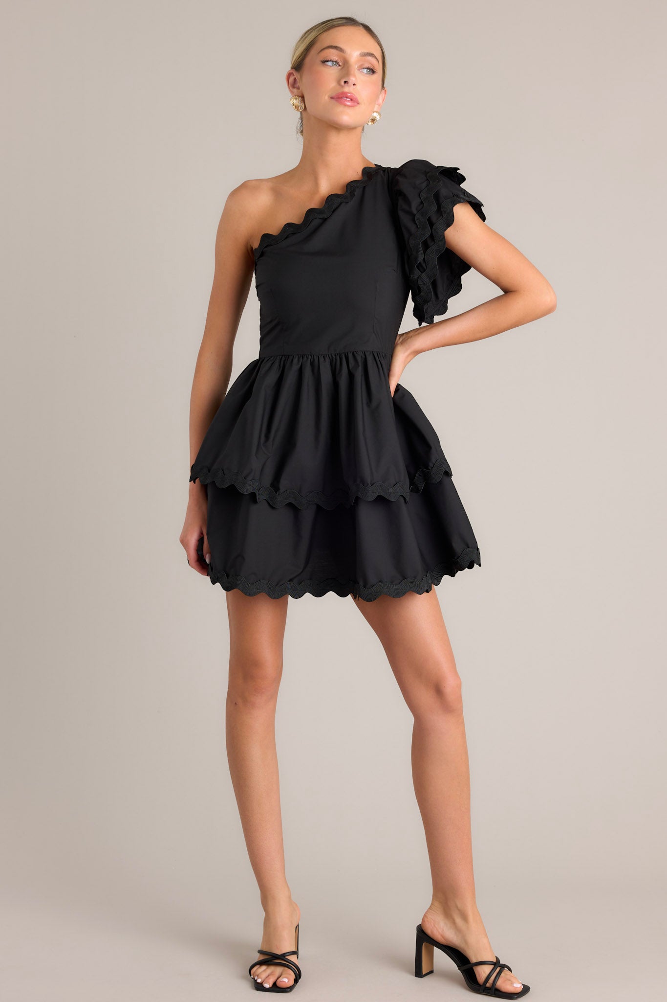 Front view of this black romper that features rick rack scalloped hems, a one shoulder neckline, a flutter sleeve, a side zipper closure, and a tiered skirt with built in shorts.
