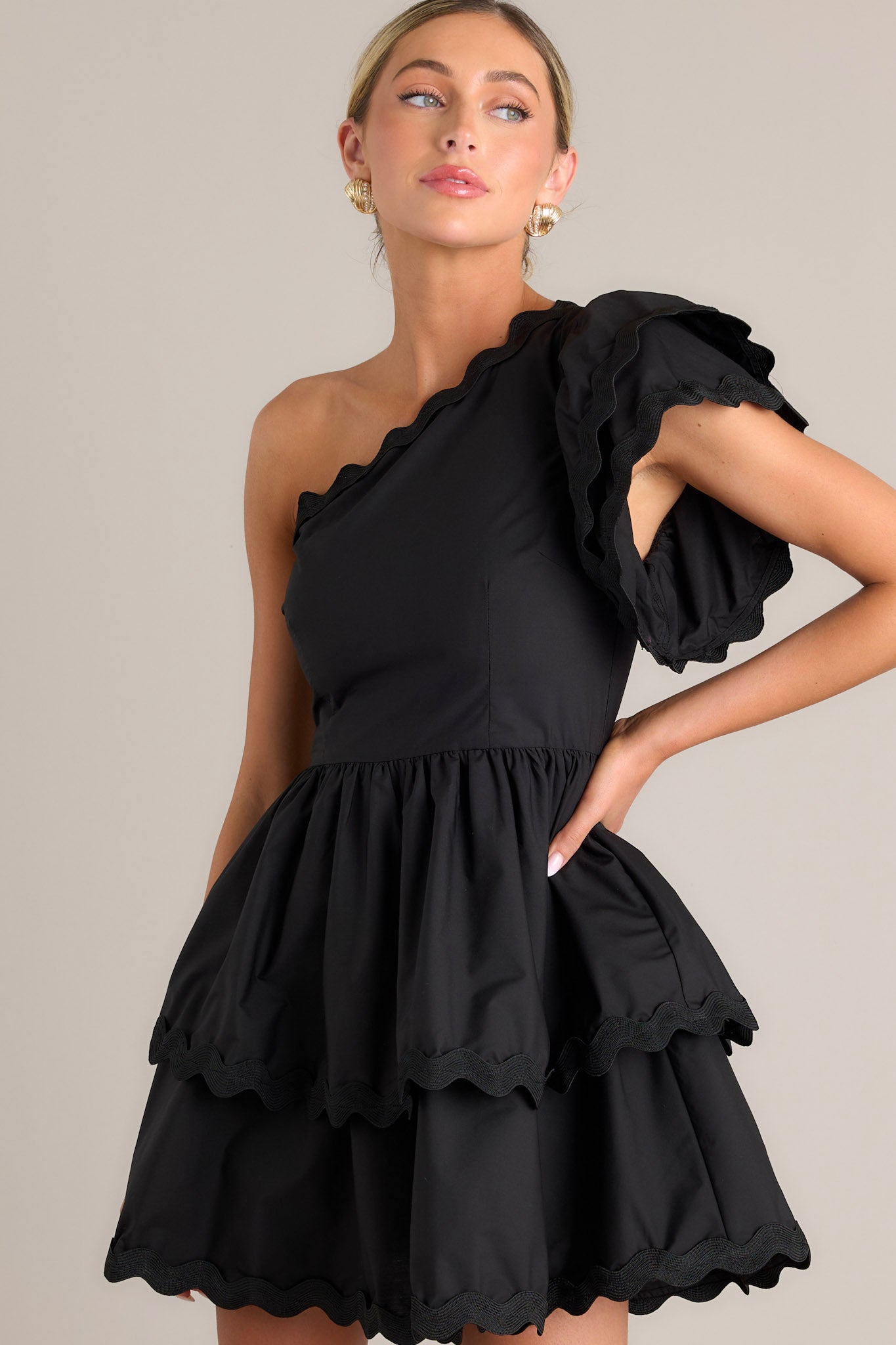 Close up front view of this black romper that features rick rack scalloped hems, a one shoulder neckline, a flutter sleeve, a side zipper closure, and a tiered skirt with built in shorts.