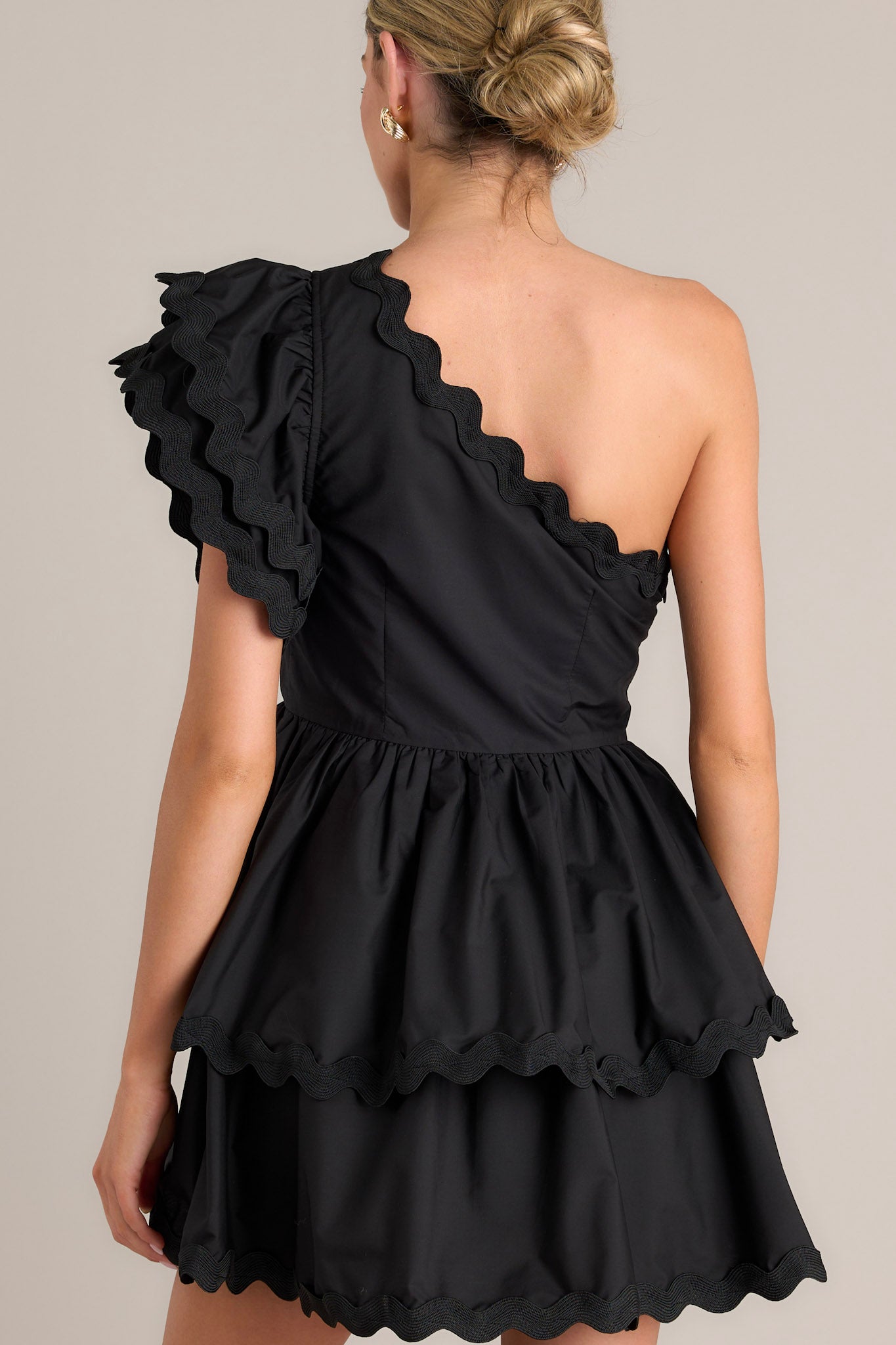 Back view of this black romper that features rick rack scalloped hems, a one shoulder neckline, a flutter sleeve, a side zipper closure, and a tiered skirt with built in shorts.
