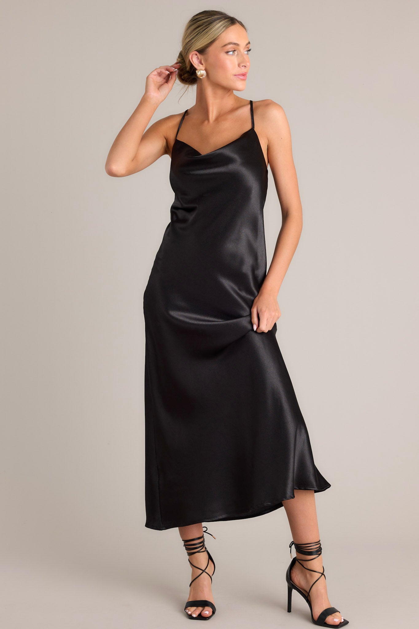 Angled front view of this elegant black dress that features a satin-like fabric, adjustable straps, and a cowl neckline